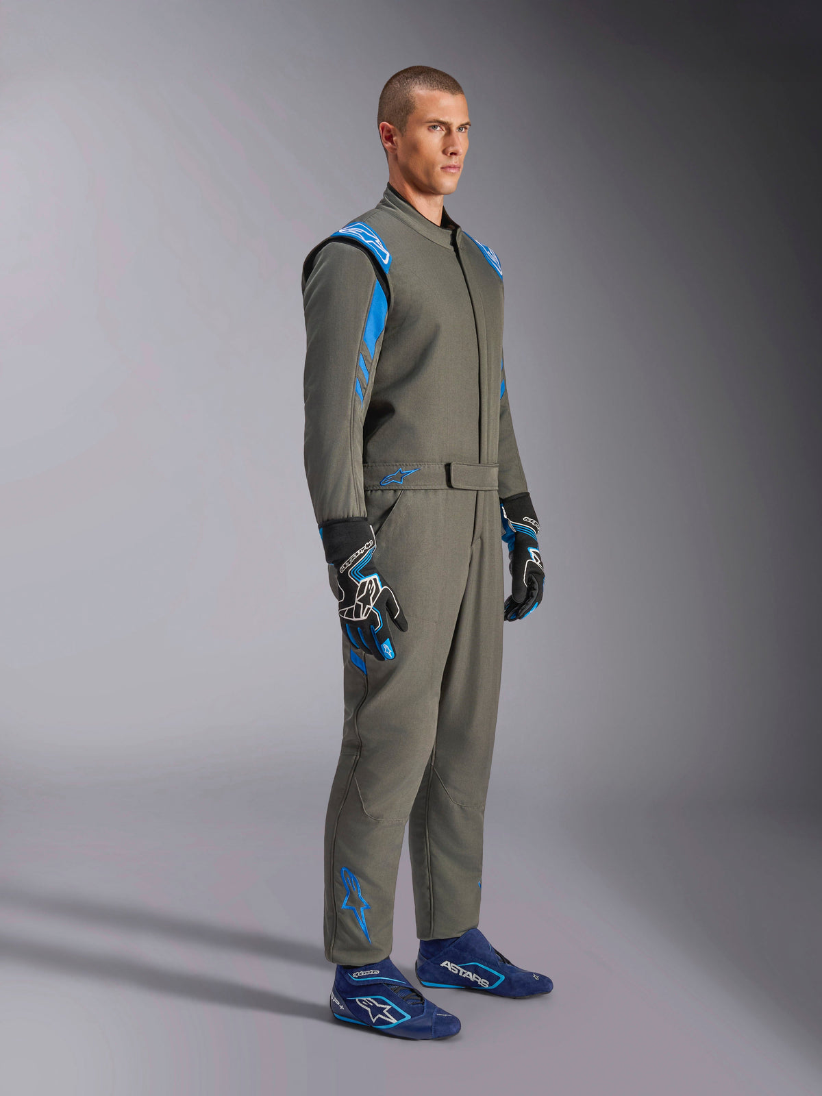 Alpinestars GP Race V3 Anzug FIA in Anthrazit-Blau, einteiliger Auto-Rennanzug getragen von einem männlichen Model, mit leuchtend blauen Akzenten an den Schultern und Knöcheln, kombiniert mit passenden Rennhandschuhen und blauen Rennschuhen, professionelle Motorsport-Ausrüstung.