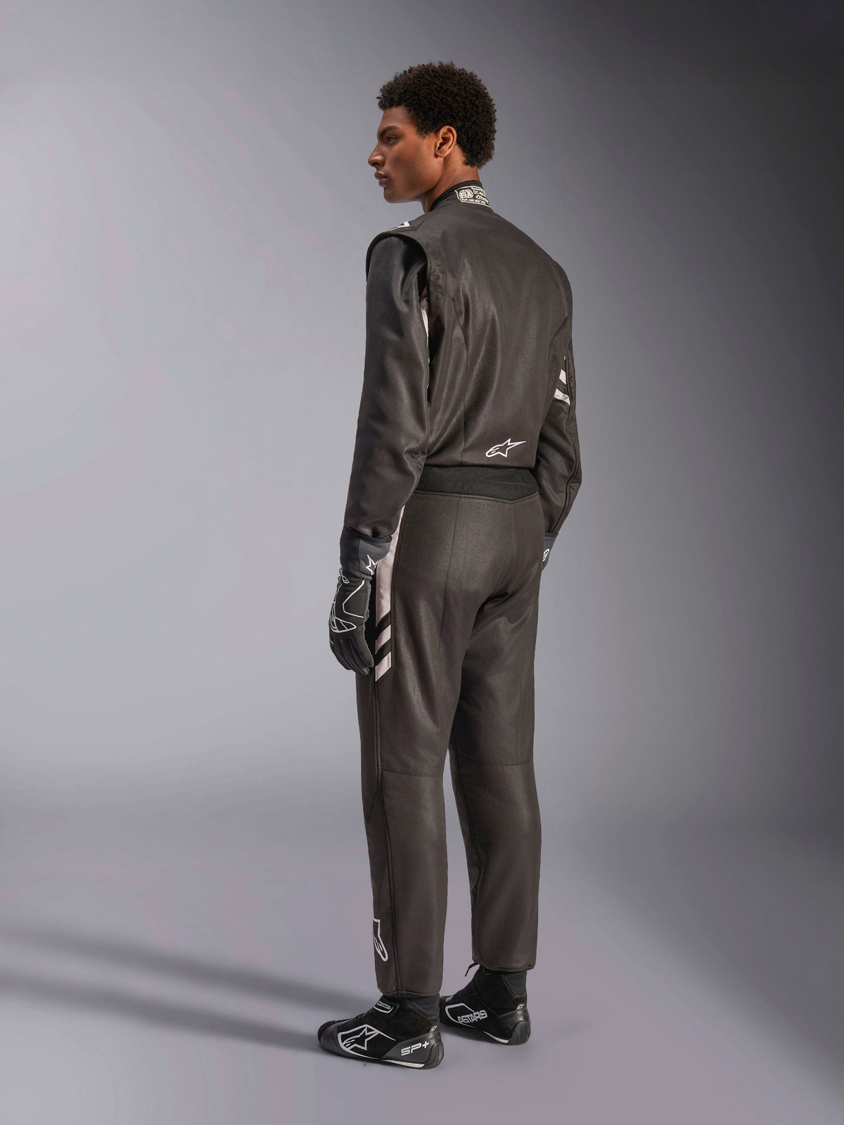 Alpinestars GP Race V3 Suit FIA, black auto racing suit with silver accents, worn by a model in a three-quarter rear view, paired with matching black racing gloves and boots, professional motorsport gear featuring a sleek two-layer design.