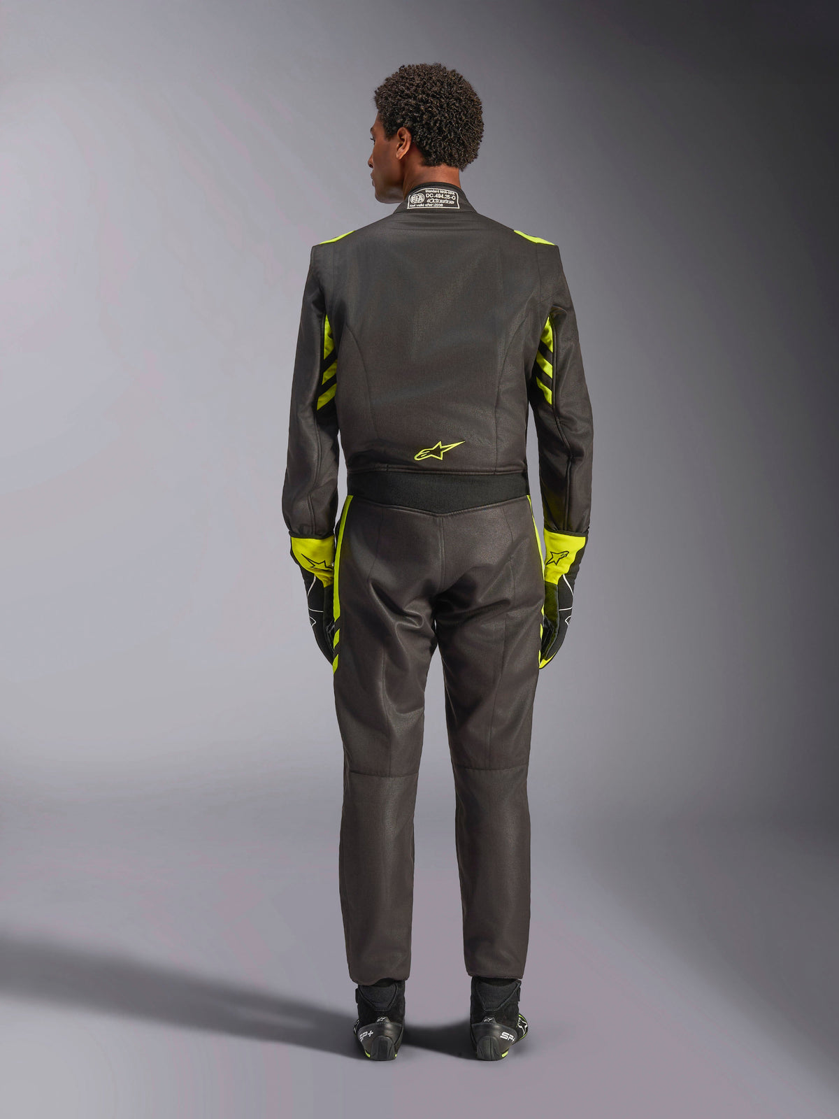 Alpinestars GP Race V3 Anzug FIA, Rückansicht, Schwarz mit gelben Fluo-Akzenten, mit gelben Einsätzen an Armen und Oberkörper, gelbes Logo am unteren Rücken, kombiniert mit passenden Rennhandschuhen und Stiefeln, professioneller Automobil-Rennanzug.