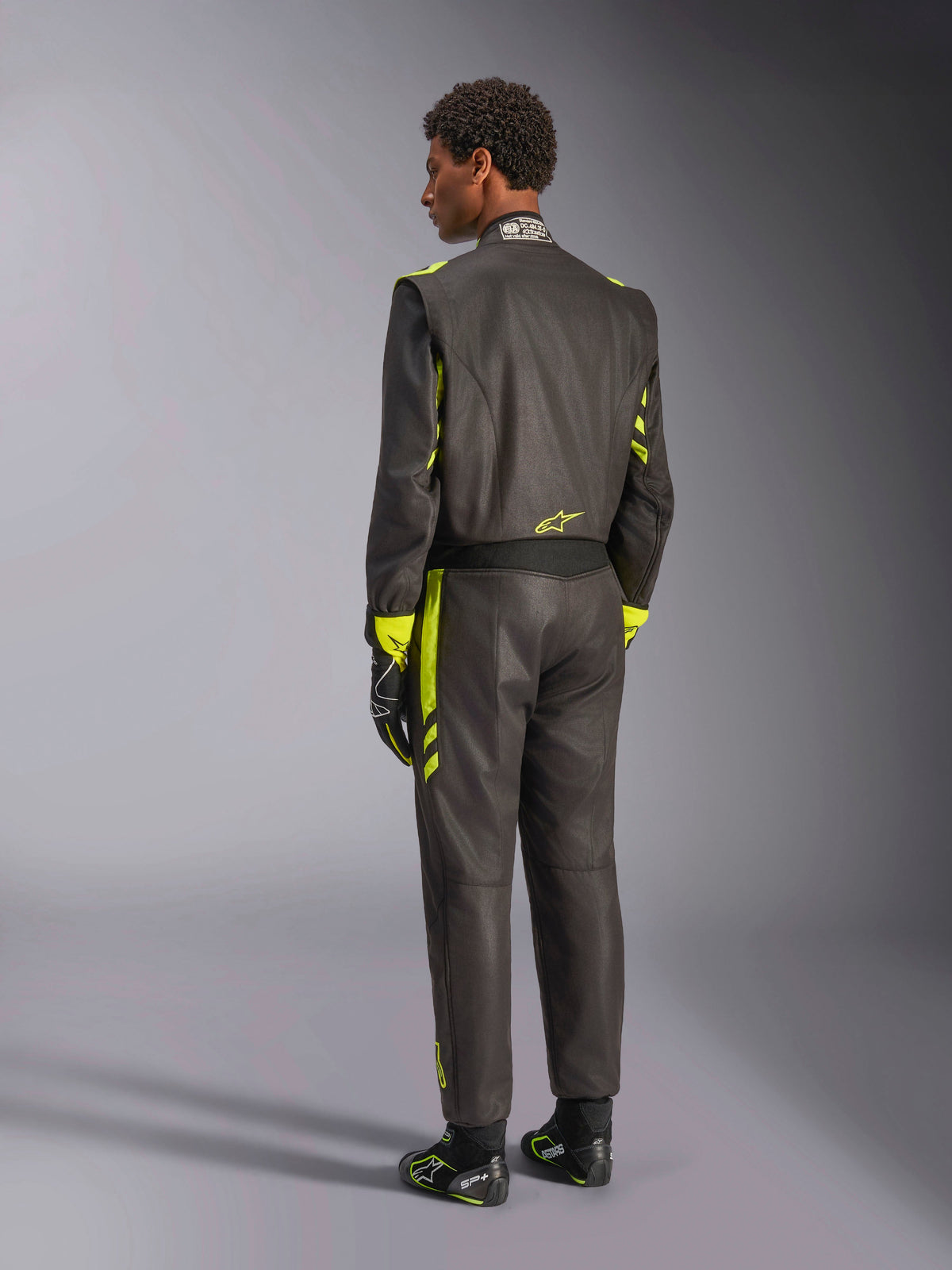 Rückansicht des Alpinestars GP Race V3 FIA Anzugs, Schwarz mit gelb-fluoreszierenden Akzenten, getragen mit passenden Rennhandschuhen und Stiefeln, professionelle Autorenn-Ausrüstung mit zweilagiger Aramid-Konstruktion