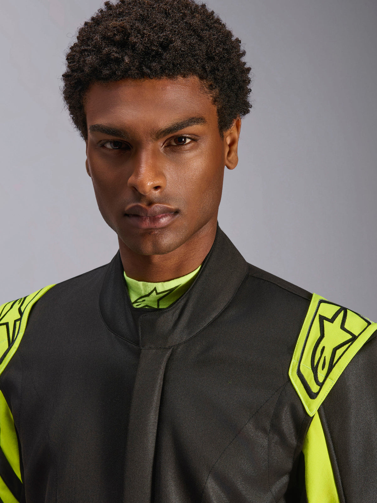 Nahaufnahme eines Models im Alpinestars GP Race V3 Kombi FIA in Schwarz und Gelb Fluo, mit gelben Extraktions-Epauletten an den Schultern mit Stern-Logos, hohem Kragendesign, professionelle Autorenn-Ausrüstung.