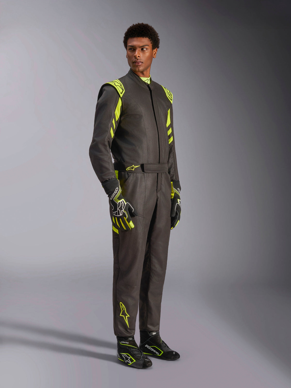 Alpinestars GP Race V3 Anzug FIA, Auto-Rennanzug, Schwarz und Fluo-Gelb, getragen von einem Model mit passenden Rennhandschuhen und Stiefeln, mit leuchtend gelben Akzenten an Schultern und Beinen, professionelle Motorsport-Ausrüstung