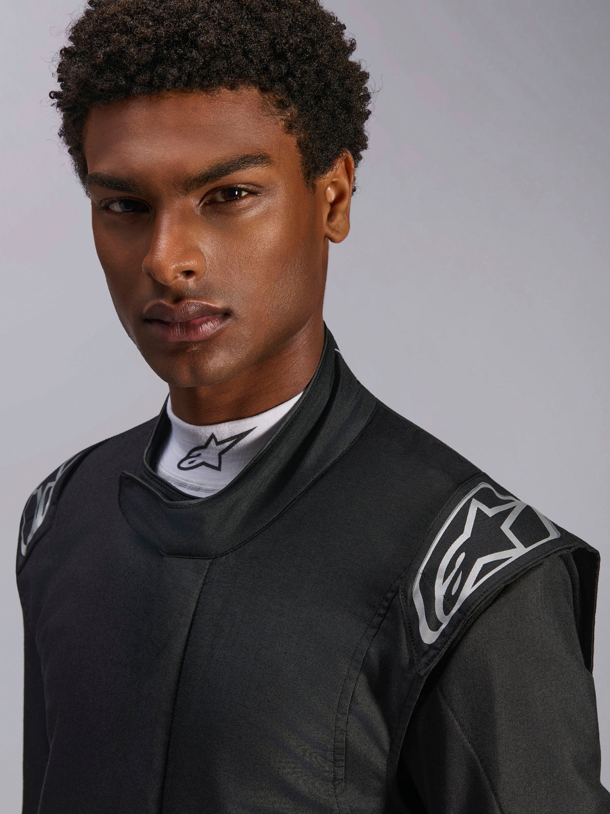 Combinaison Alpinestars Hypertech V3, combinaison de course automobile Noir Dark Shadow, gros plan sur un mannequin homme, logos argentés sur les épaules, col montant avec sous-pull blanc Alpinestars, équipement de protection professionnel de sport automobile