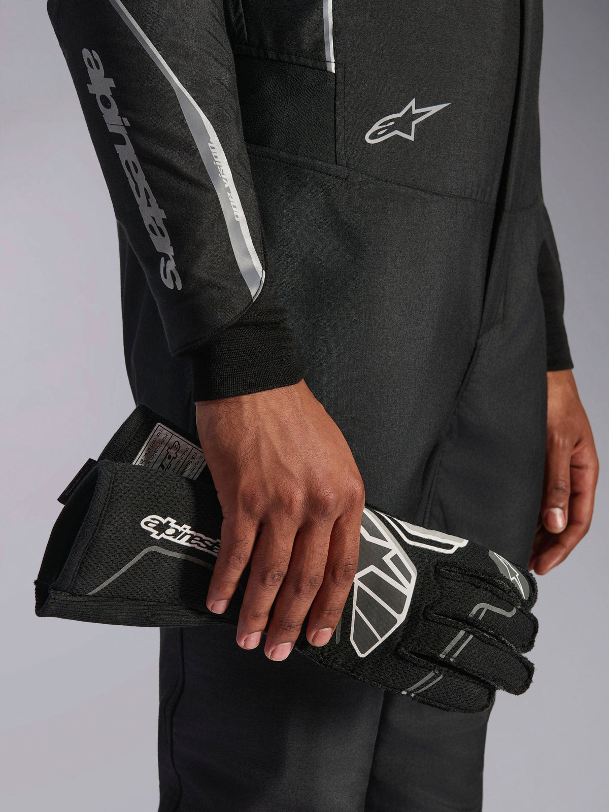 Combinaison Alpinestars Hypertech V3 en Noir Dark Shadow, gros plan sur les détails de la manche et du torse avec marquage Alpinestars argenté, main tenant un gant de course noir avec des accents blancs, équipement de course automobile professionnel