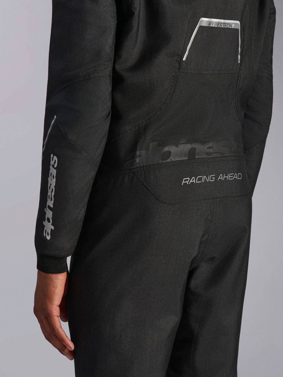 Combinaison Alpinestars Hypertech V3 en Noir/Dark Shadow, vue rapprochée montrant les graphismes à l'arrière 'RACING AHEAD' et 'HYPERTECH V3', logo blanc sur la manche, équipement de course automobile professionnel.