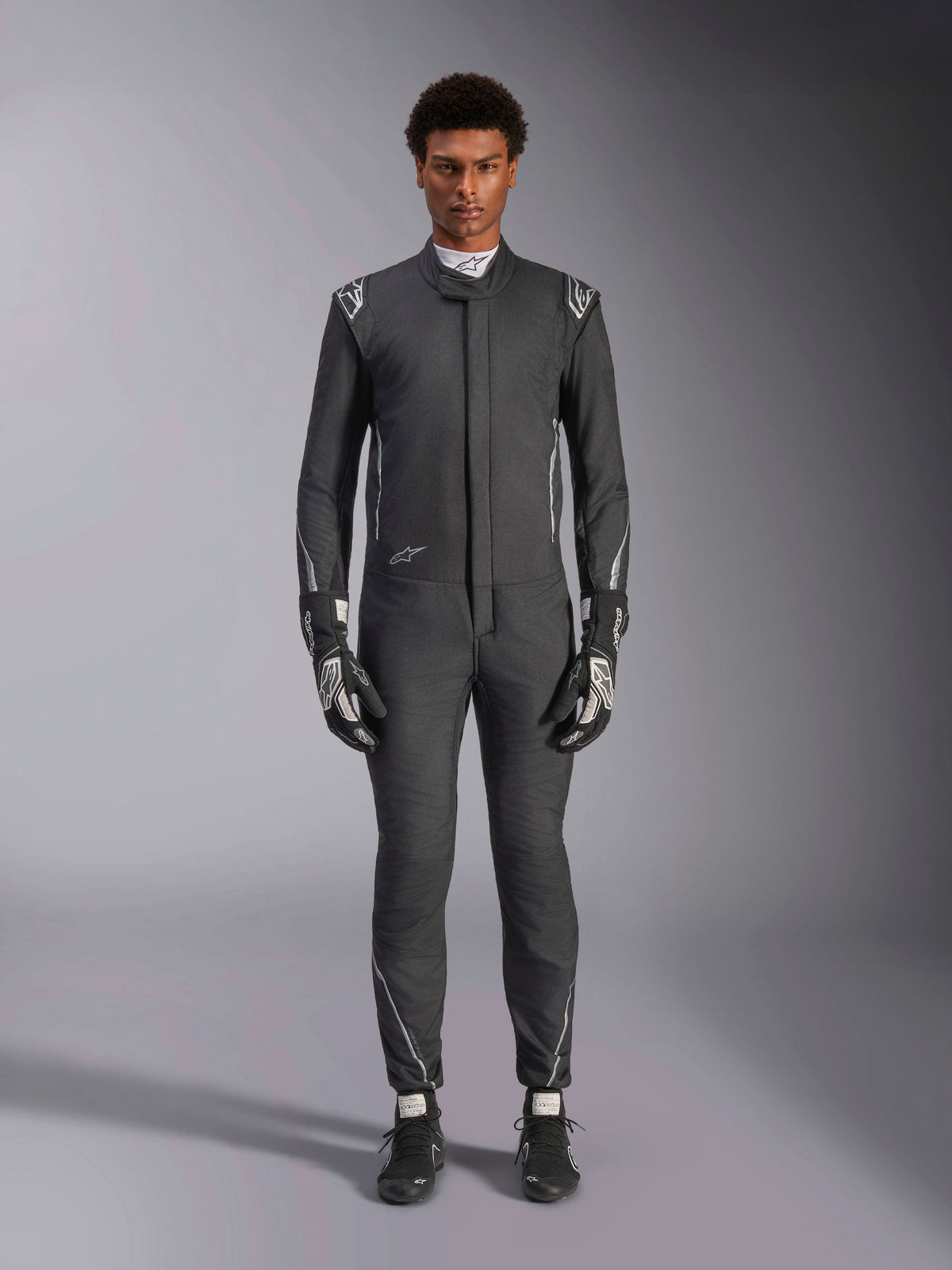 Combinaison Alpinestars Hypertech V3, combinaison de course automobile professionnelle, Noir et Dark Shadow, portée par un mannequin avec gants et chaussures de course assortis, coupe athlétique slim avec accents argentés et logos, équipement de sport automobile haute performance
