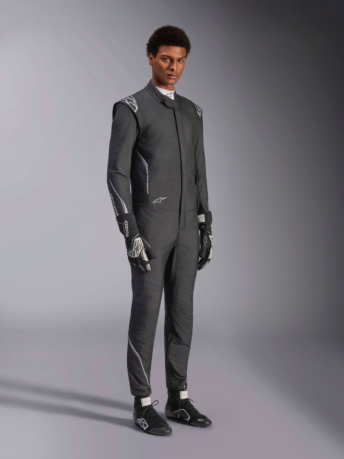 Combinaison Alpinestars Hypertech V3, combinaison de course automobile, noir et Dark Shadow, portée par un mannequin homme avec gants noirs et chaussures de course assortis, coupe athlétique slim, logos Alpinestars et accents gris clair, équipement de protection professionnel pour sport automobile