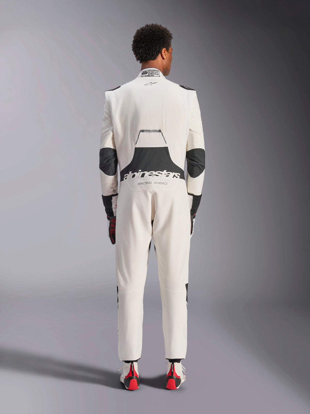 Achteraanzicht van een model dat het Alpinestars Hypertech V3-pak in White Black Silver draagt, met het grote zwarte lumbale stretchpaneel met 'alpinestars' en 'RACING AHEAD' branding, zwarte ellebooginzetstukken, FIA-certificeringslabel op de kraag en een atletische, slanke pasvorm ontworpen voor professionele racerij.