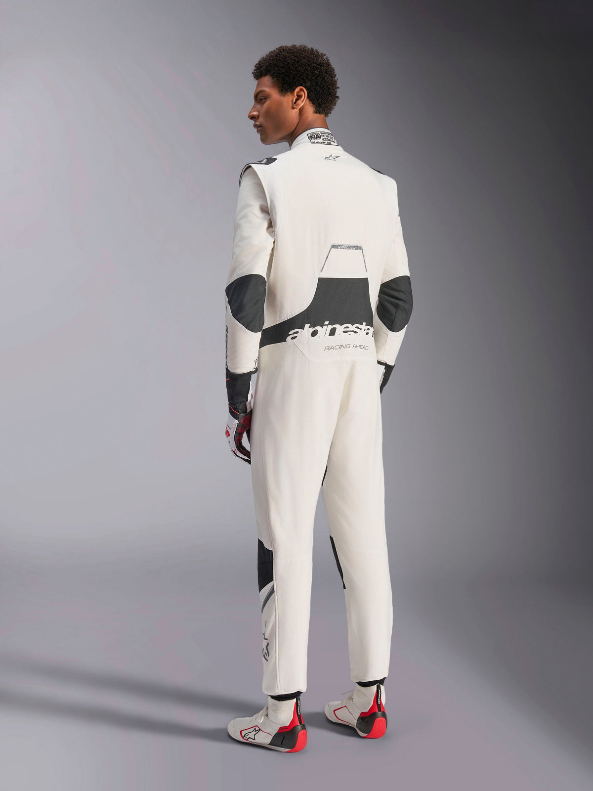 Alpinestars Hypertech V3-pak, professioneel autoracepak, wit, zwart en zilver, achteraanzicht van model met zwarte stretchpanelen op de lendenen en ellebogen, Alpinestars-branding op de rug, gecombineerd met bijpassende racehandschoenen en laarzen voor een complete motorsport-look