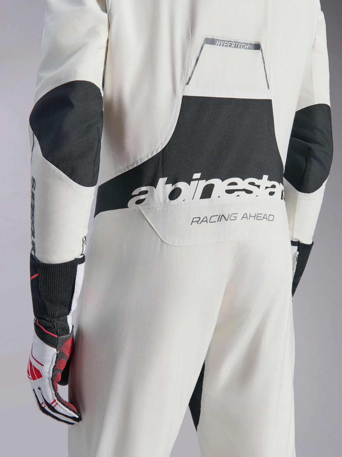 Alpinestars Hypertech V3-pak, wit, zwart en zilveren autoracepak, achteraanzicht met groot zwart 'alpinestars'-logo en 'RACING AHEAD'-tekst, voorzien van zwarte stretchpanelen op de ellebogen en het lendegebied, professionele motorsport beschermende kleding.