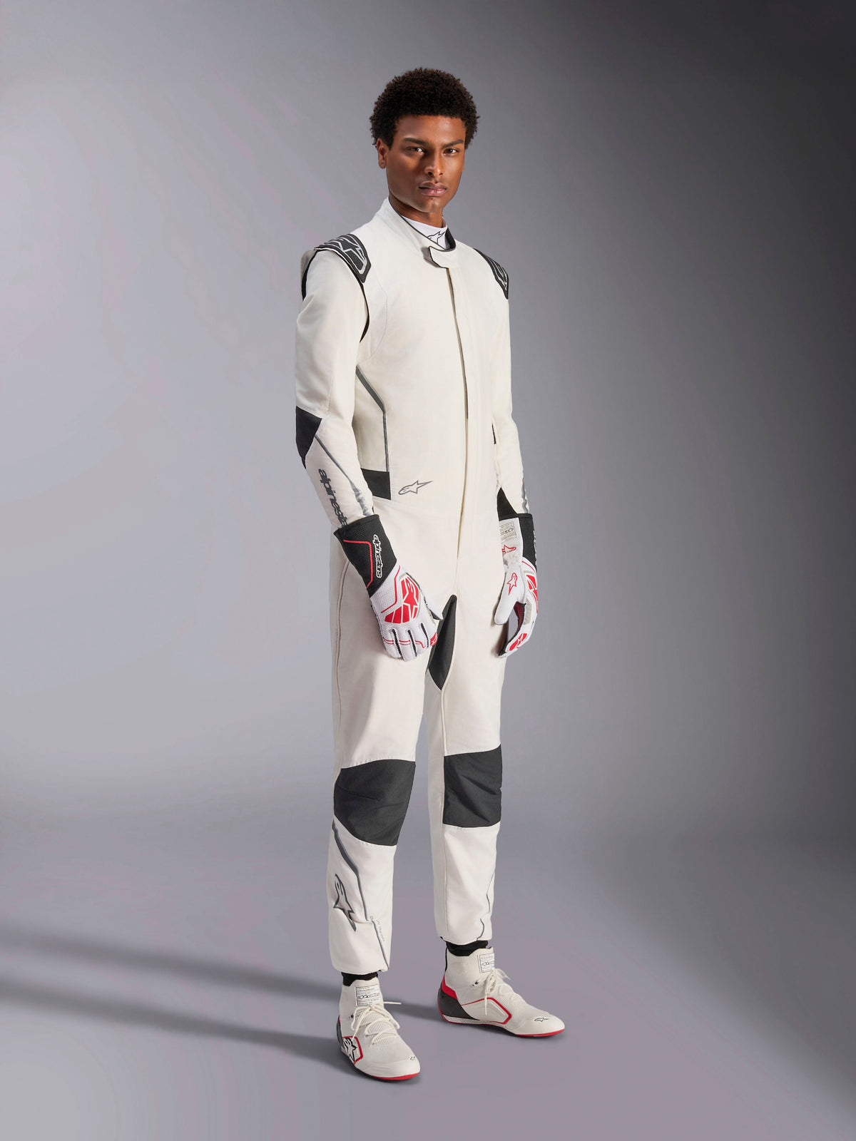 Mannelijk model draagt het Alpinestars Hypertech V3-pak, wit met zwarte en zilveren accenten, professionele autorace-uitrusting met zwarte panelen op de schouders en knieën, gestyled met wit-rode racehandschoenen en witte racelaarzen