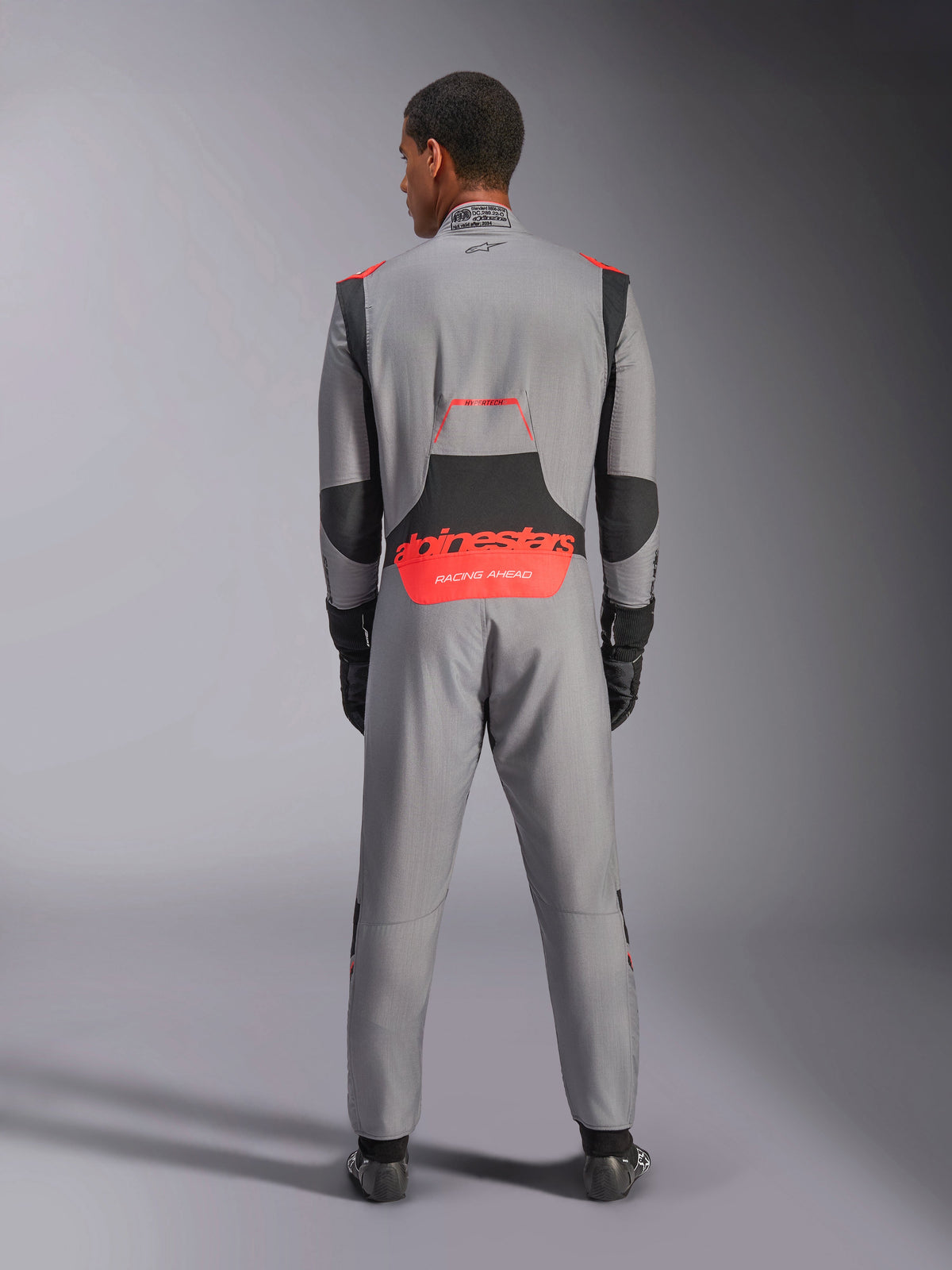 Alpinestars Hypertech V3 Anzug, professioneller Autorennanzug, Mittelgrau mit schwarzen und fluoreszierend roten Akzenten, Rückansicht an einem Model mit rotem Alpinestars-Branding am unteren Rücken, leichte Nomex-Konstruktion für Performance auf der Rennstrecke.