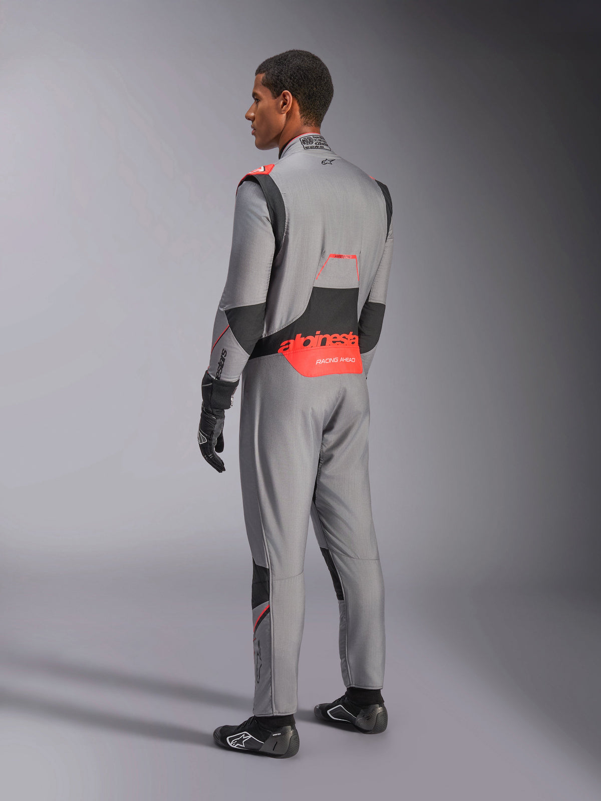 Alpinestars Hypertech V3 Anzug in Mid Gray Black Red Fluo, Dreiviertel-Rückansicht, mit markantem rotem Alpinestars-Branding am unteren Rücken, schwarzen Stretcheinsätzen und einer schlanken, athletischen Passform für den professionellen Rennsport.