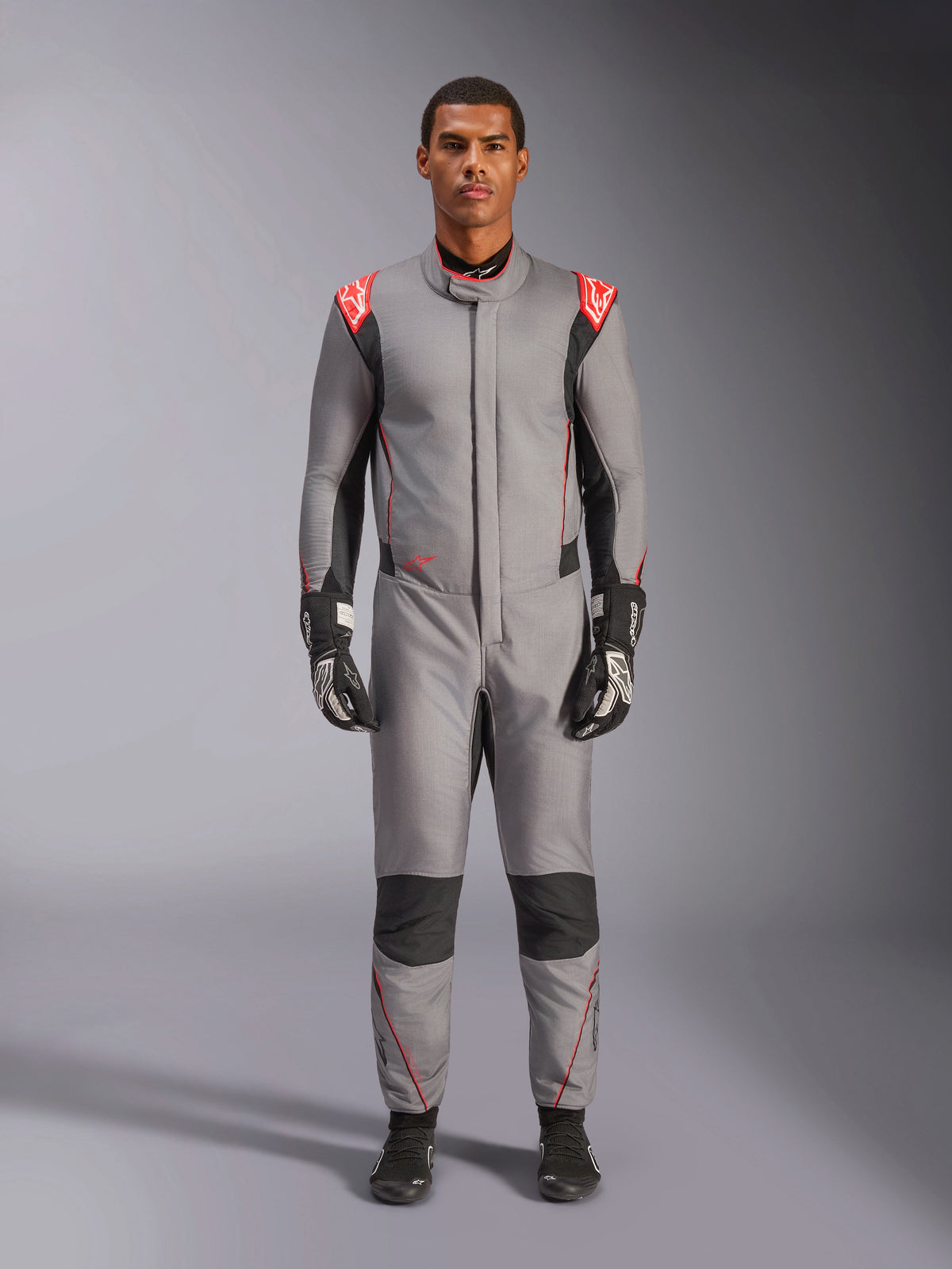 Alpinestars Hypertech V3 Anzug, professioneller Autorennanzug in Mittelgrau, Schwarz und Leuchtrot, getragen von einem Model mit schwarzen Rennhandschuhen und Schuhen, athletisch-schlanke Passform, leuchtrote Schulterakzente, schwarze Einsätze an den Knien und Seiten