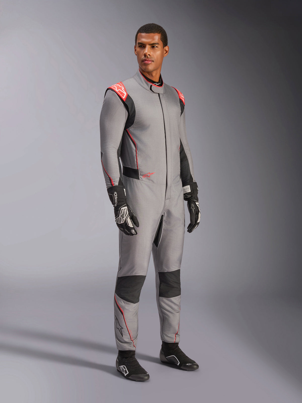 Männliches Model im Alpinestars Hypertech V3 Anzug in Mittelgrau mit schwarzen und neonroten Akzenten, professioneller Autorennanzug kombiniert mit schwarzen Rennhandschuhen und -schuhen, leichte zweilagige Nomex-Konstruktion mit roten Schulterdetails und athletischer Passform