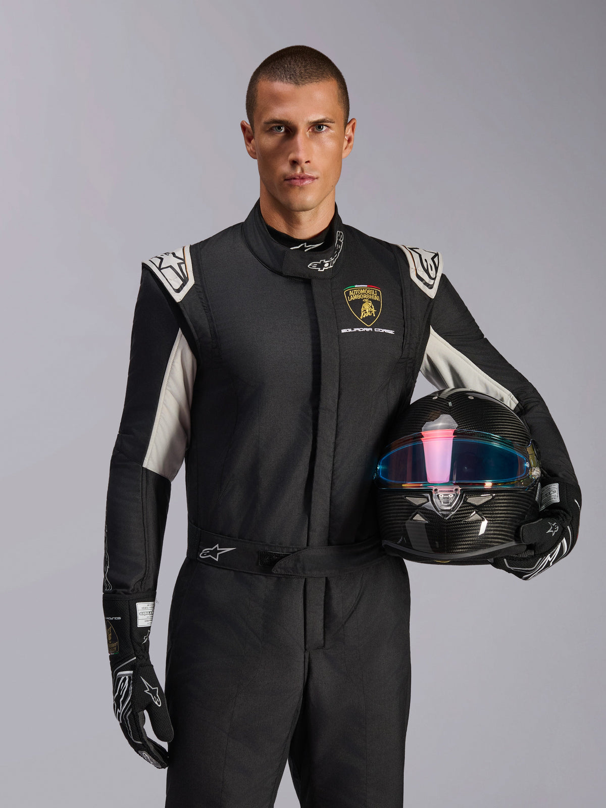 Modèle masculin portant la combinaison Limited Edition Lamborghini GP Tech V4, combinaison de course noire avec accents argentés et logo Lamborghini Squadra Corse, tenant un casque en fibre de carbone, gants de course noirs avec logos blancs, équipement de course automobile professionnel