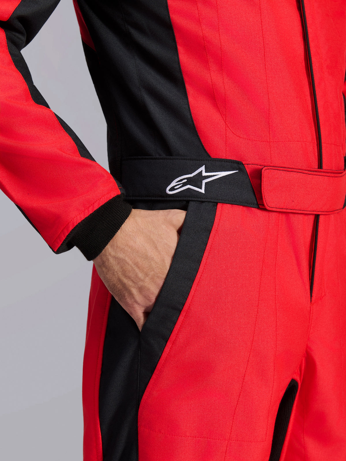 Alpinestars GP Pro Comp V2 FIA Rennanzug, Rot und Schwarz, Nahaufnahme des Taillenbereichs mit einer Hand in der Tasche, schwarzer Gürtel mit weißem Stern-Logo, strukturierte Einsätze für den Autorennsport