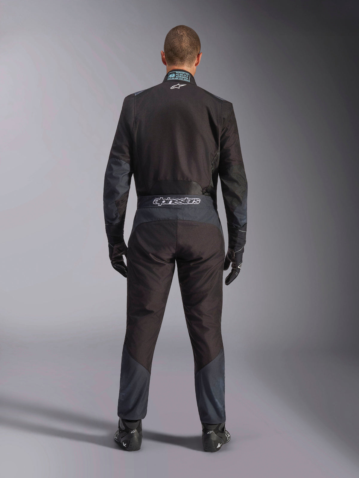 Combinaison Alpinestars KMX-5 V3, combinaison de karting, noir et Dark Shadow, vue arrière d'un pilote portant la tenue de course complète avec gants et chaussures assortis, logo Alpinestars sur la ceinture et empiècements ergonomiques.