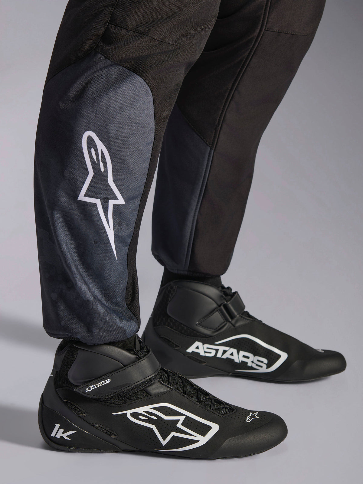 Combinaison Alpinestars KMX-5 V3, Black Dark Shadow, gros plan sur le bas des jambes et les chevilles, logo étoile blanc sur le mollet, associée à des chaussures de karting noires, mettant en avant les chevilles tricotées et la coupe athlétique-regular.