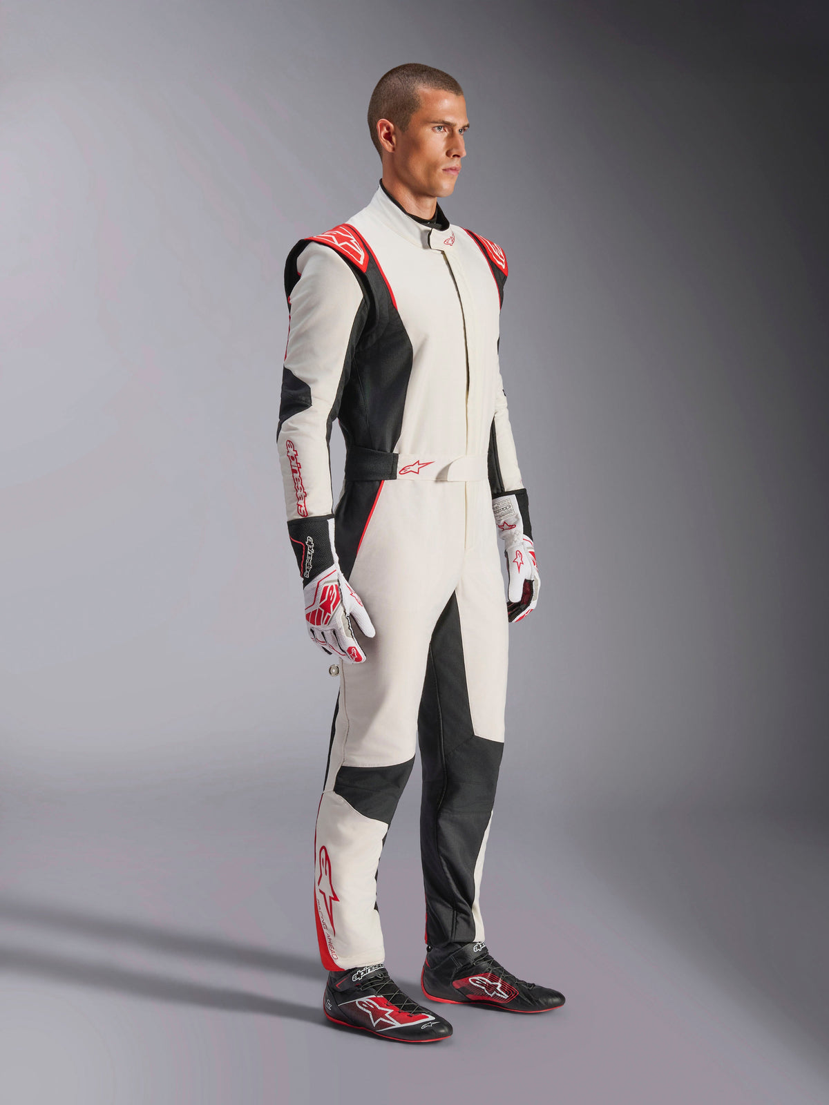 Pilote de course automobile portant une combinaison Alpinestars GP Tech V4, blanc, noir et rouge, combinaison intégrale une pièce, gants et chaussures de course assortis, équipement de sport automobile professionnel avec logo Alpinestars et design ergonomique