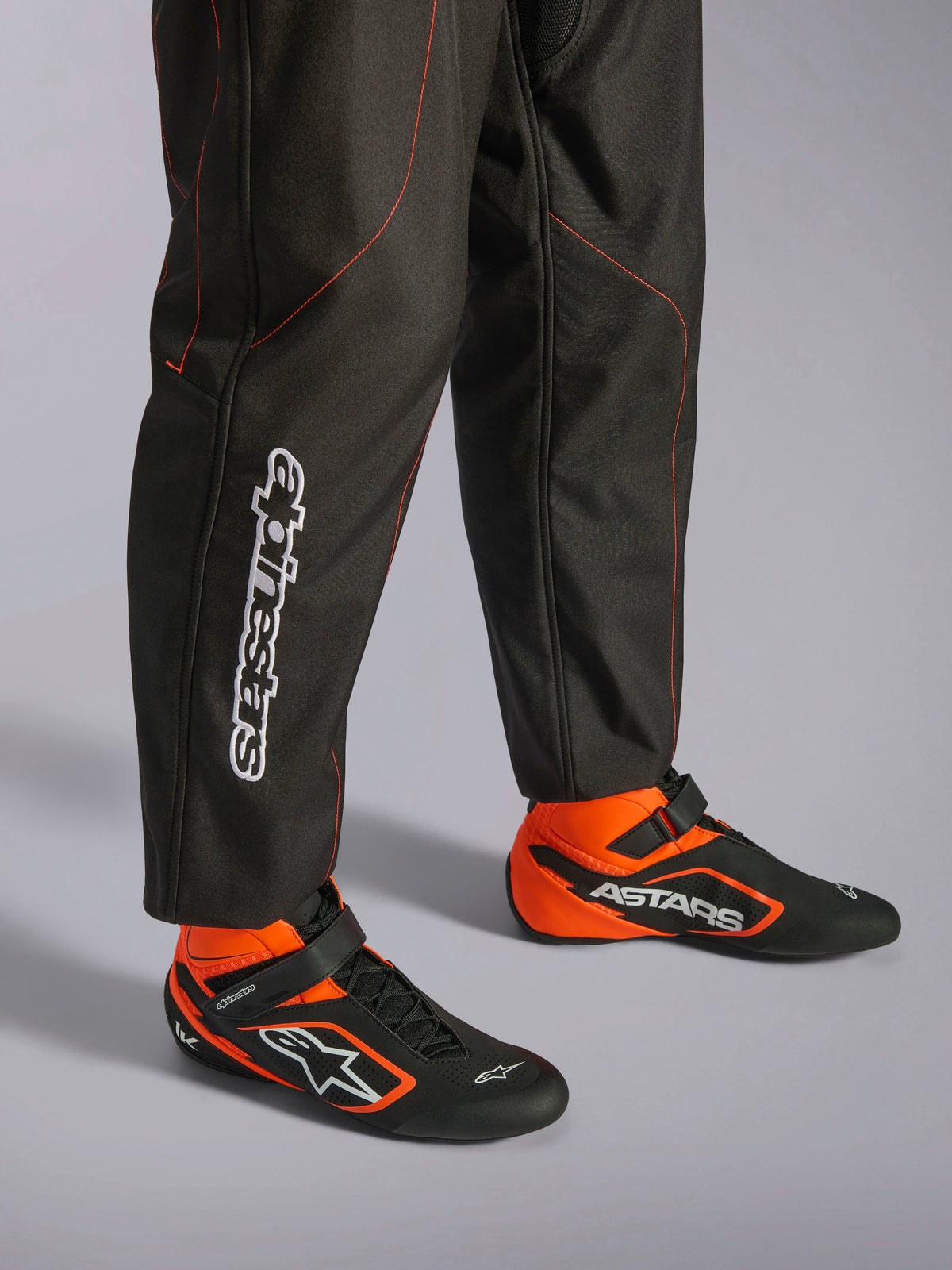 Alpinestars KMX-9 V3 Anzug, Karting-Anzug, Schwarz und Rot Fluo, Detailansicht der Unterschenkel und Rennschuhe, schwarzer Stoff mit neonroten Nähten, vertikales Alpinestars-Logo an der Wade, kombiniert mit passenden schwarz-orangenen Rennstiefeln, entwickelt für professionellen Kartsport