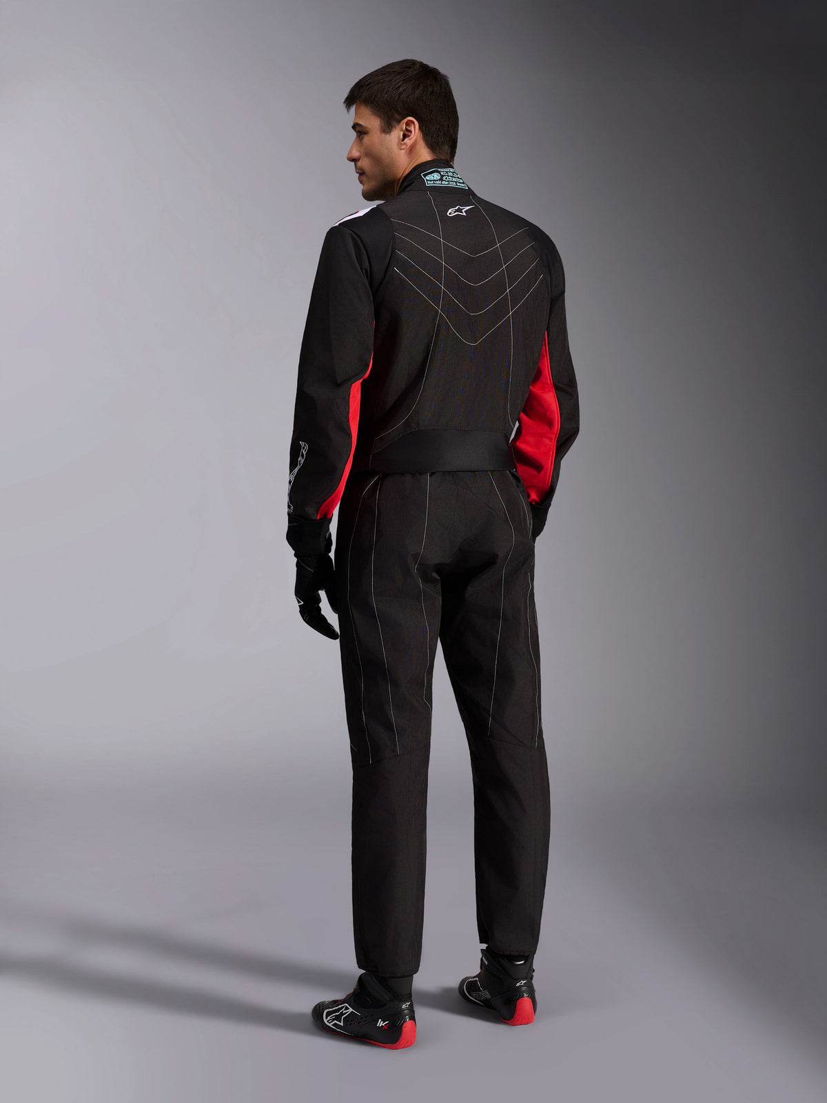 Alpinestars KMX-9 V3-pak, zwart rood wit, eendelig kartpak vanaf de achterkant gezien, met witte geometrische stiksels op de rug en rode accenten op de mouwen, gedragen met zwarte racehandschoenen en kartschoenen.
