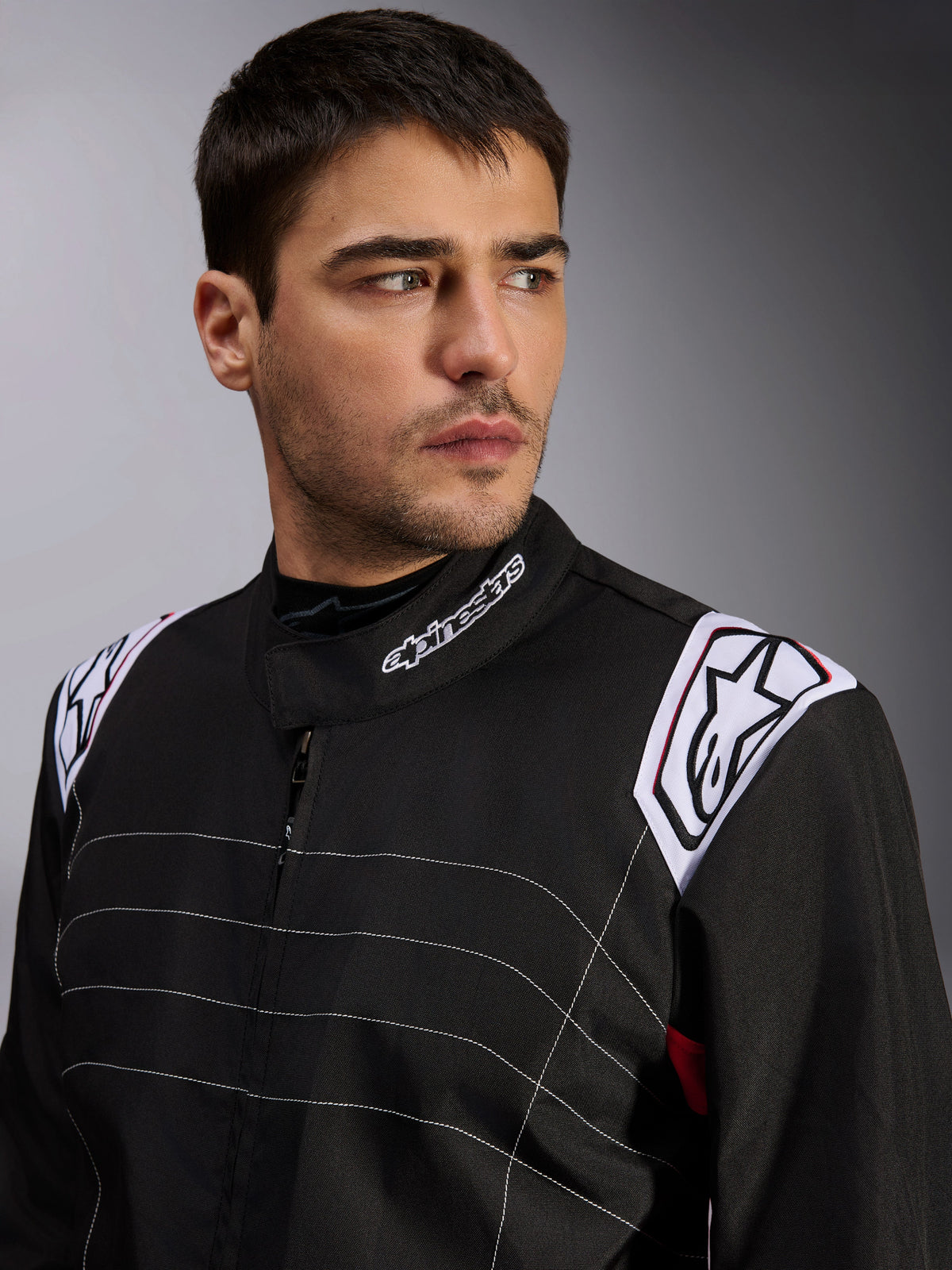 Alpinestars KMX-9 V3-pak, zwart kartpak gedragen door een mannelijk model, close-up van borst en schouders met witte branding op de kraag en epauletten, voorzien van wit decoratief stiksel en een professioneel autosportontwerp