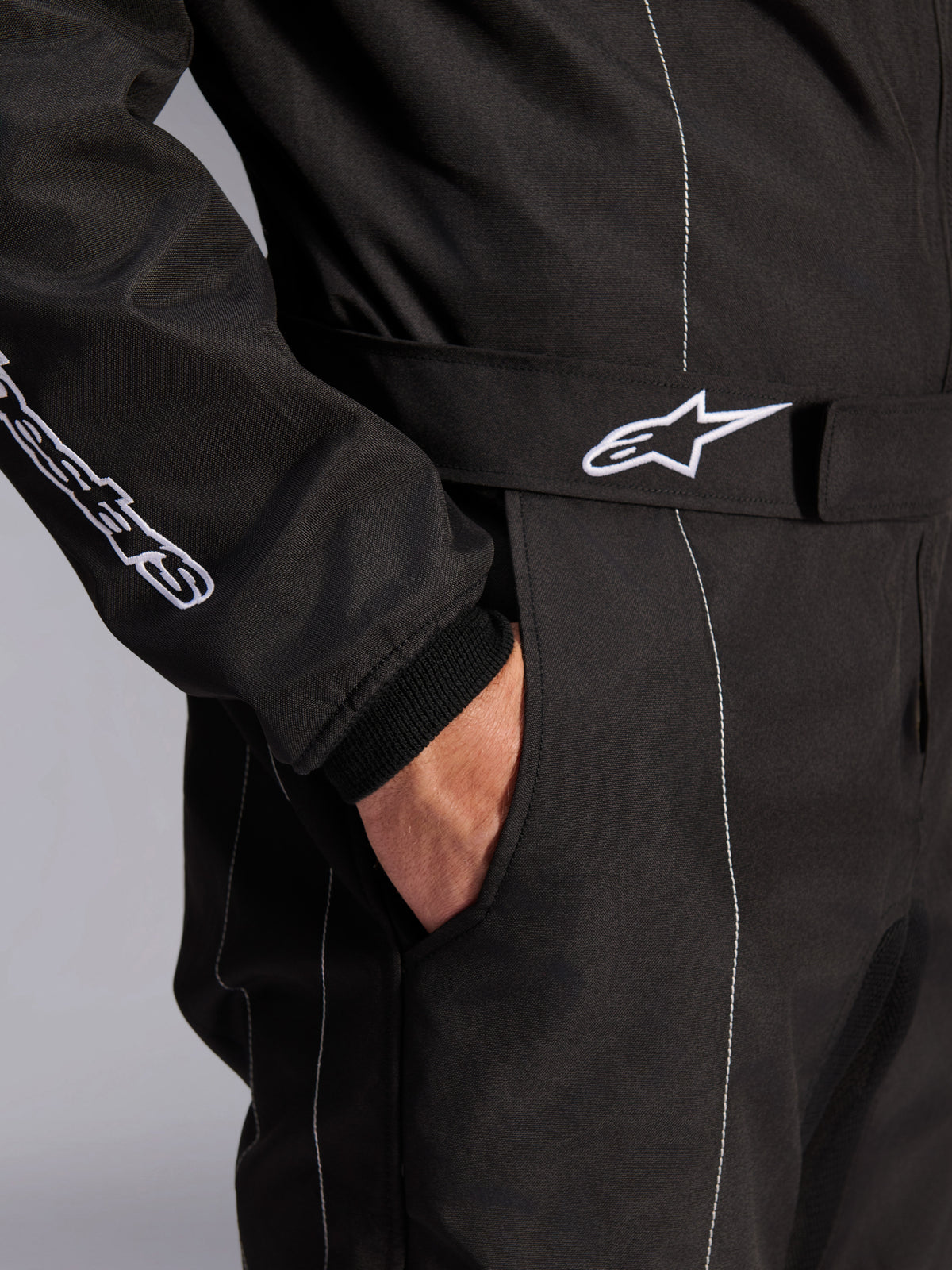 Alpinestars KMX-9 V3 Anzug, Schwarz-Weiß, Nahaufnahme des verstellbaren Taillengürtels mit besticktem Logo, Seitentasche, weiße Kontrastnähte auf schwarzem Stoff, technisches Detail des Karting-Anzugs