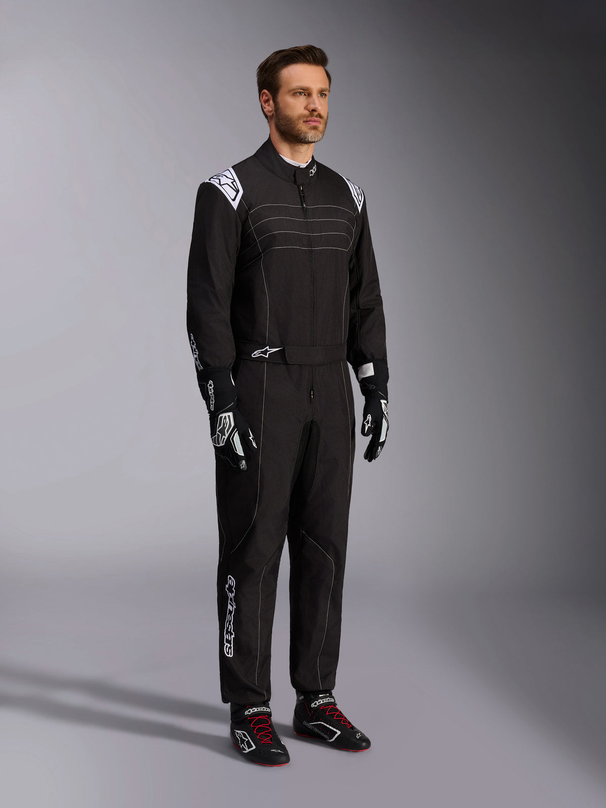 Alpinestars KMX-9 V3 Karting-Anzug, Schwarz und Weiß, männliches Model in vollständigem Rennanzug mit weißem Alpinestars-Logo auf den Epauletten, kombiniert mit schwarz-weißen Rennhandschuhen und schwarzen Kart-Schuhen mit roten Schnürsenkeln, Studioaufnahme zeigt Front- und Seitenprofil