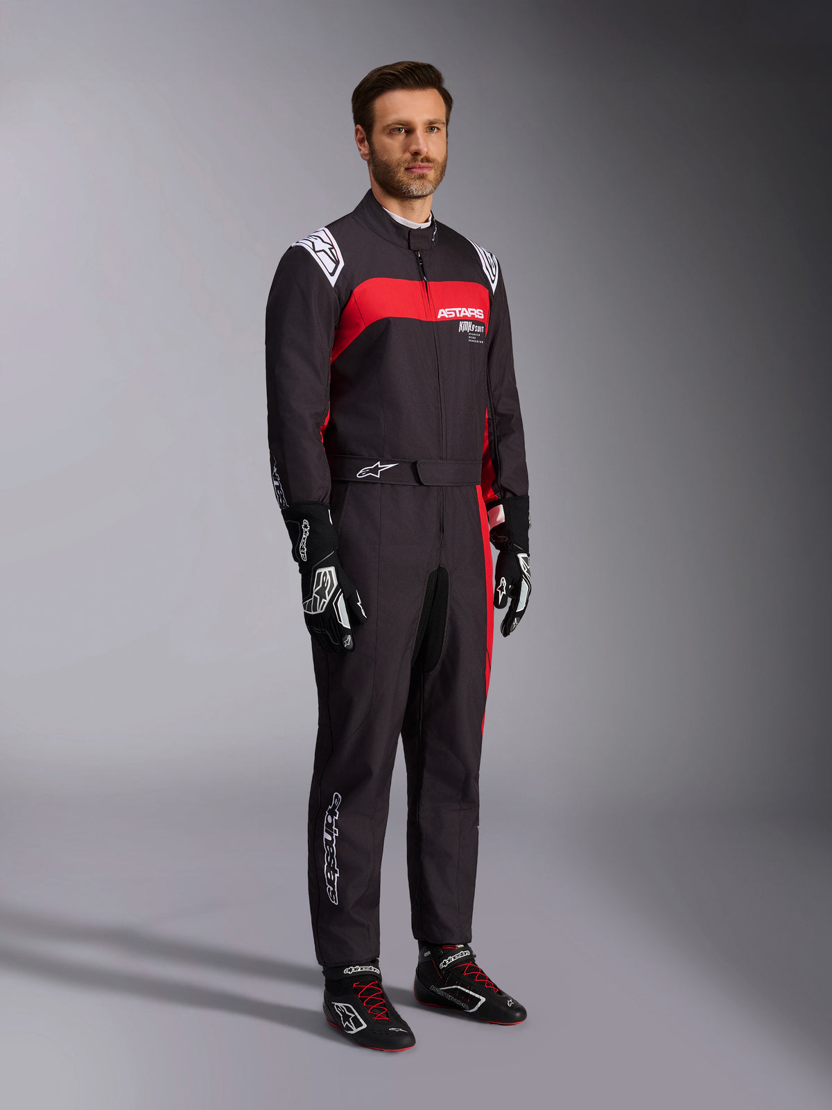 Male model wearing Alpinestars KMX-9 V3 Graphic 3 Suit, Karting Suit, Black and Red, featuring white shoulder epaulettes and red chest branding, paired with black racing gloves and shoes with red laces, professional motorsport apparel.