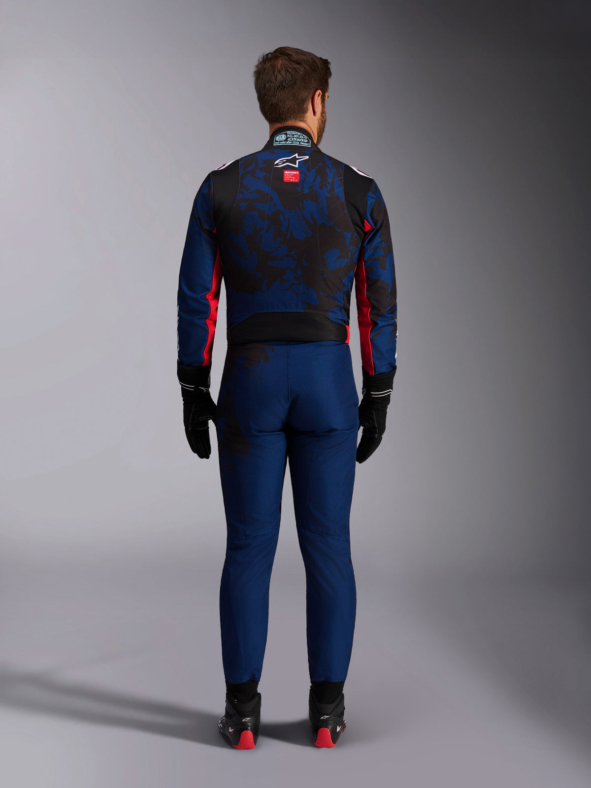 Pilote de karting vu de dos portant la combinaison KMX-9 V3 Graphic 5 en Navy Red, présentant un motif graphique bleu marine et noir, des accents latéraux rouges et le logo Alpinestars sur le haut du dos, associée à des gants et des chaussures de course noirs.