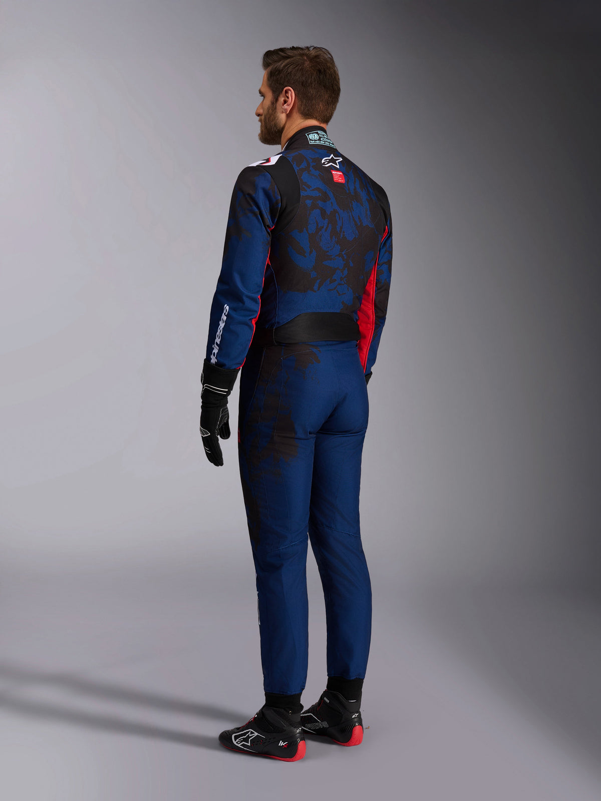 Alpinestars KMX-9 V3 Graphic 5 Suit, combinaison de karting, bleu marine et rouge, vue arrière montrant un corps bleu marine avec un motif graphique sombre d'inspiration camouflage sur le dos et des accents rouges, portée avec des gants de course noirs et des chaussures de karting noires et rouges.