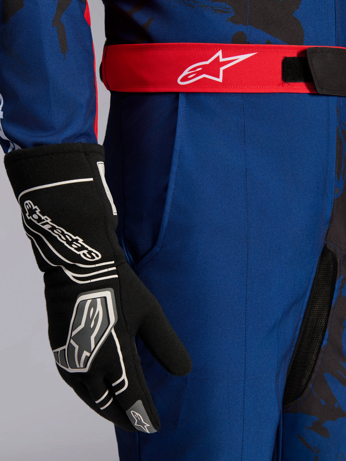 Combinaison Alpinestars KMX-9 V3 Graphic 5, combinaison de karting, bleu marine et rouge, gros plan montrant la ceinture réglable rouge avec logo blanc, portée avec un gant de karting noir, tissu bleu marine résistant et poche de cuisse intégrée