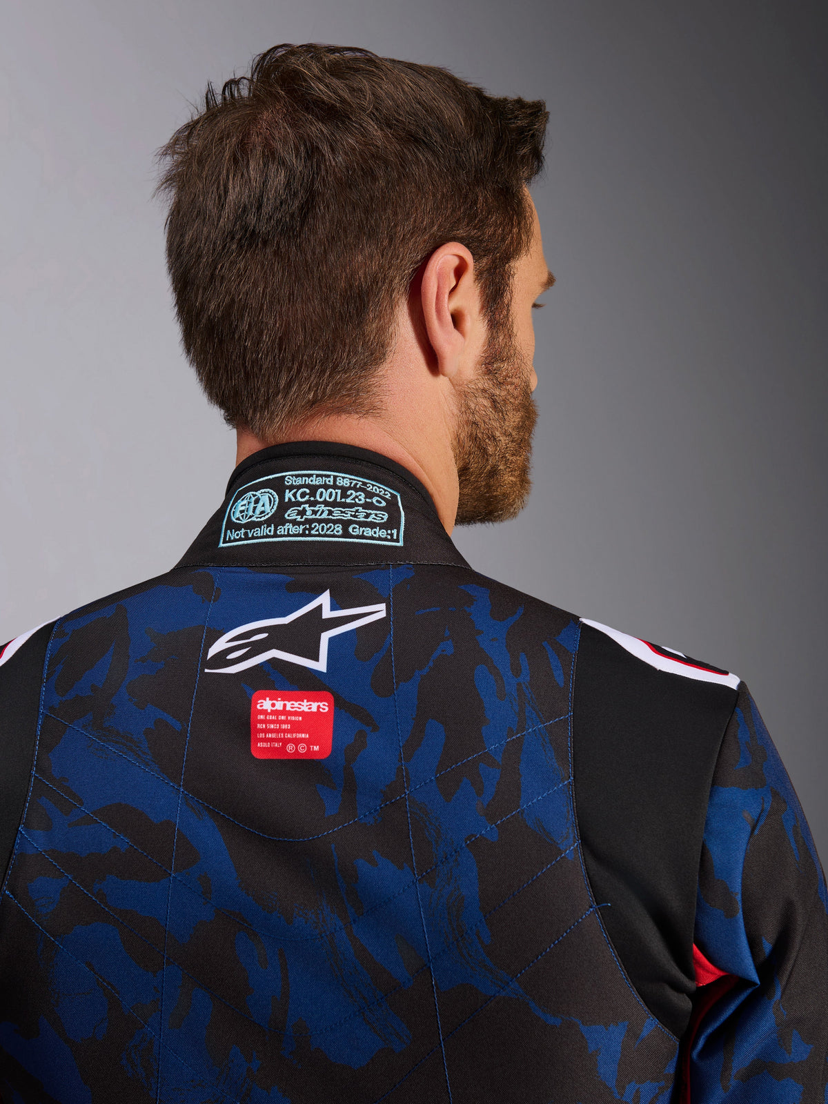 Gros plan de la combinaison Alpinestars KMX-9 V3 Graphic 5, combinaison de karting bleu marine et rouge, présentant un motif camouflage bleu et noir, un logo étoile blanc et une étiquette de marque rouge sur le haut du dos, avec un patch de certification FIA 8877-2022 sur le col