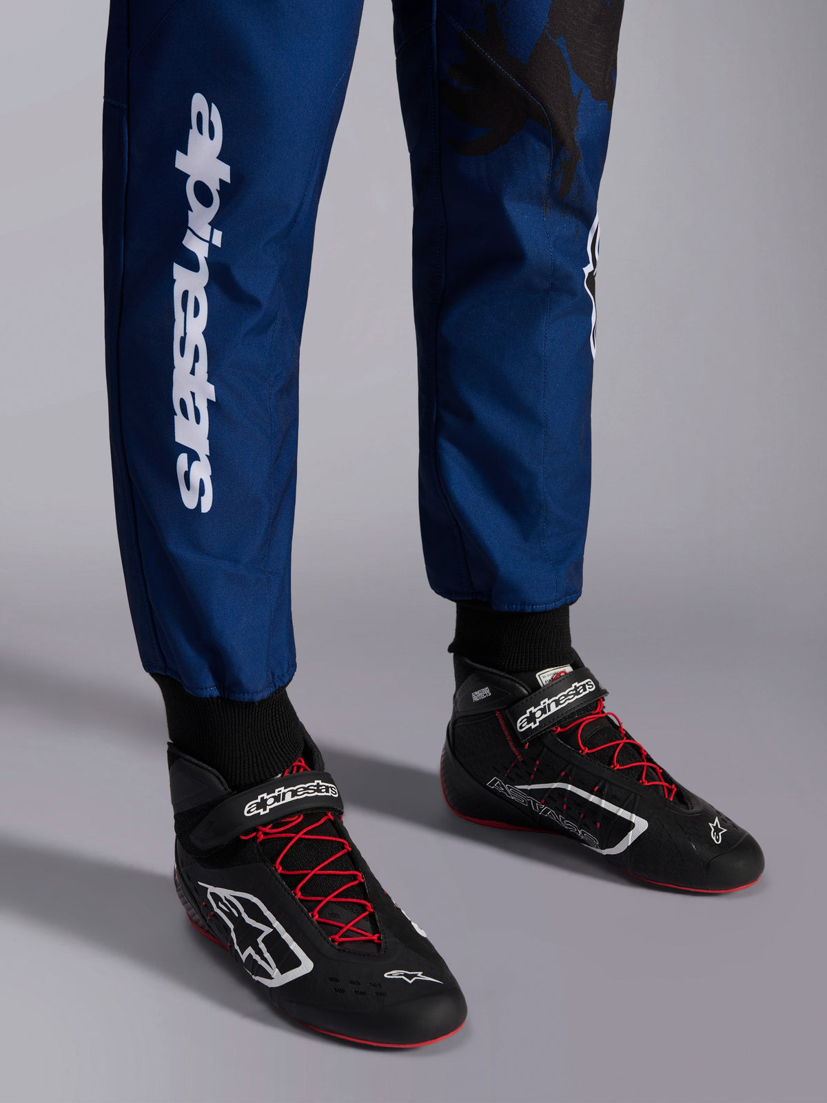 Combinaison Alpinestars KMX-9 V3 Graphic 5, bleu marine et rouge, gros plan sur le bas des jambes et les chaussures de karting, tissu bleu marine avec marquage vertical Alpinestars blanc, chaussures noires avec lacets rouges et logos blancs, équipement de karting professionnel