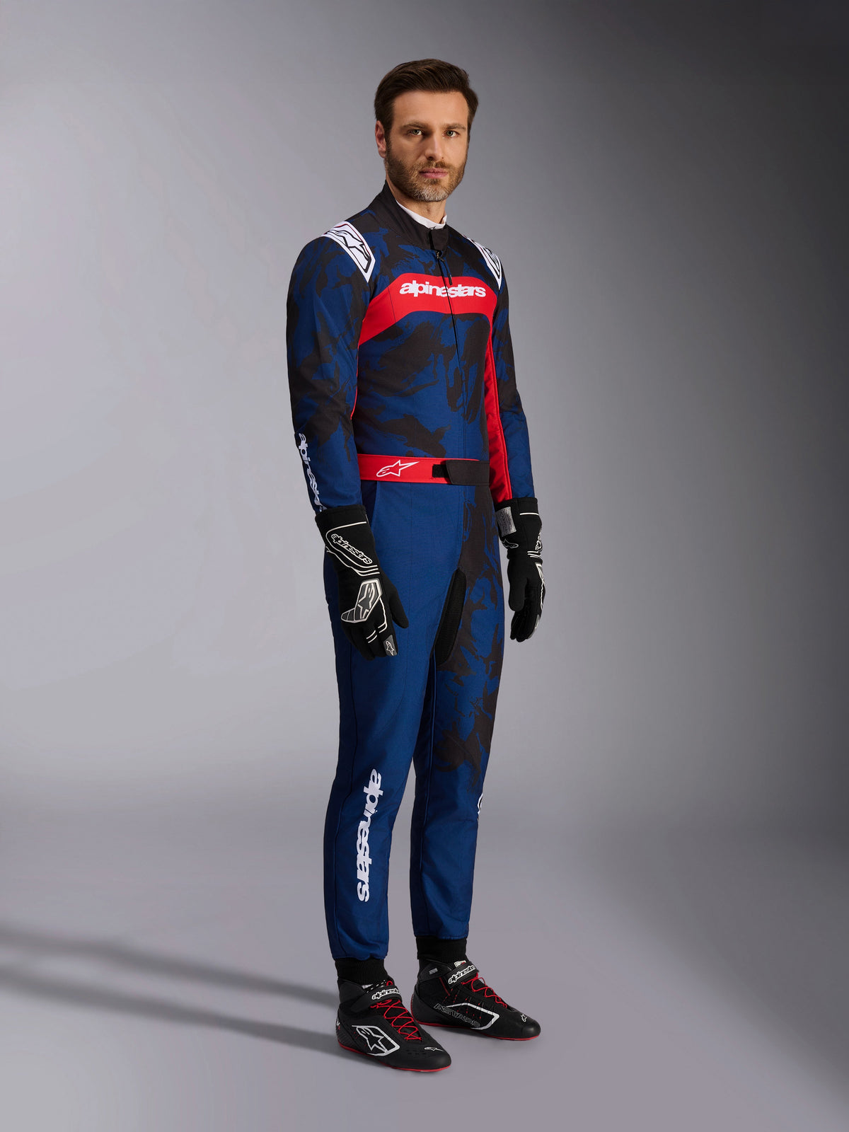 Modèle portant la combinaison Alpinestars KMX-9 V3 Graphic 5 Navy Red, vue en pied avec gants de karting et chaussures de course noirs, combinaison bleu marine aux motifs camouflage sombres et empiècement rouge sur la poitrine, équipement de protection professionnel pour le karting