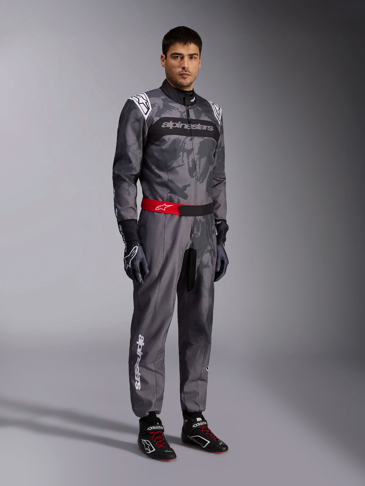 Alpinestars KMX-9 V3 Graphic 5 Anzug, Kart-Anzug, Dunkelgrau und Schwarz mit Camouflage-Grafikmuster, roter Taillengürtel mit weißem Logo, getragen mit Rennhandschuhen und Rennschuhen, professionelle Motorsport-Ausrüstung für den Kartsport