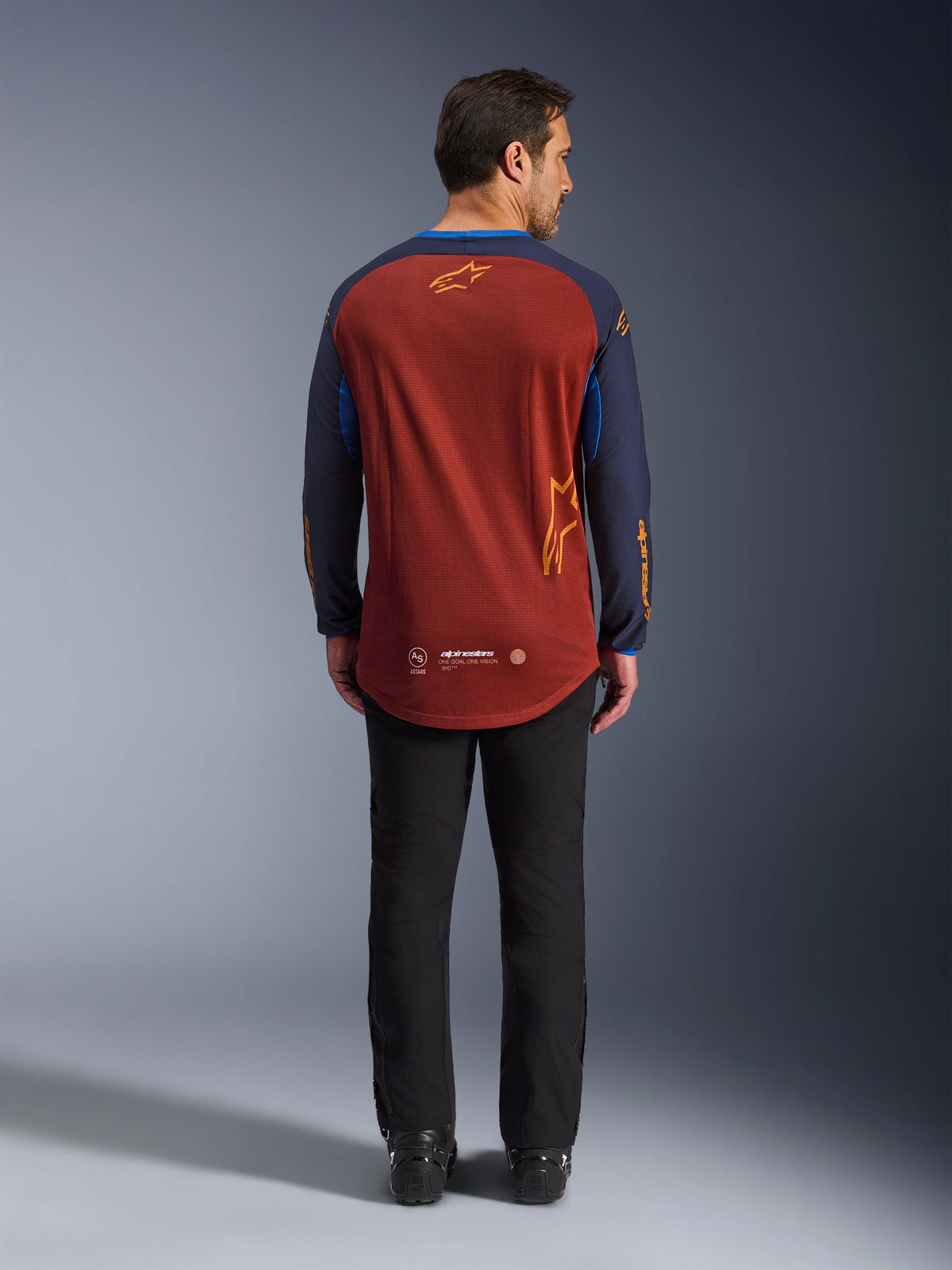 Maillot Alpinestars Maxdura Dual en Rust Brown et Mood Indigo, vue de dos montrant l'ourlet arrière allongé et les détails du logo, porté avec un pantalon technique noir et des bottes, conçu pour l'aventure et la moto dual-sport.