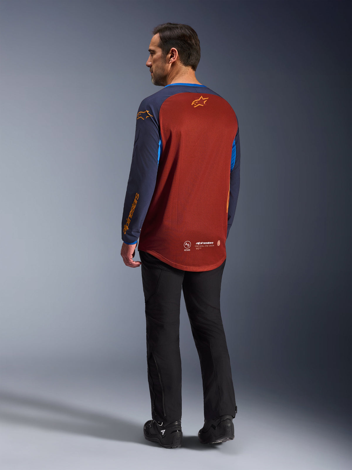 Maillot Alpinestars Maxdura Dual, rouille et bleu Mood Indigo, maillot de moto d'aventure vu de dos, panneau en maille de couleur rouille pour la ventilation, manches bleu marine avec logos orange, porté avec un pantalon de pilotage et des bottes noirs, équipement technique tout-terrain
