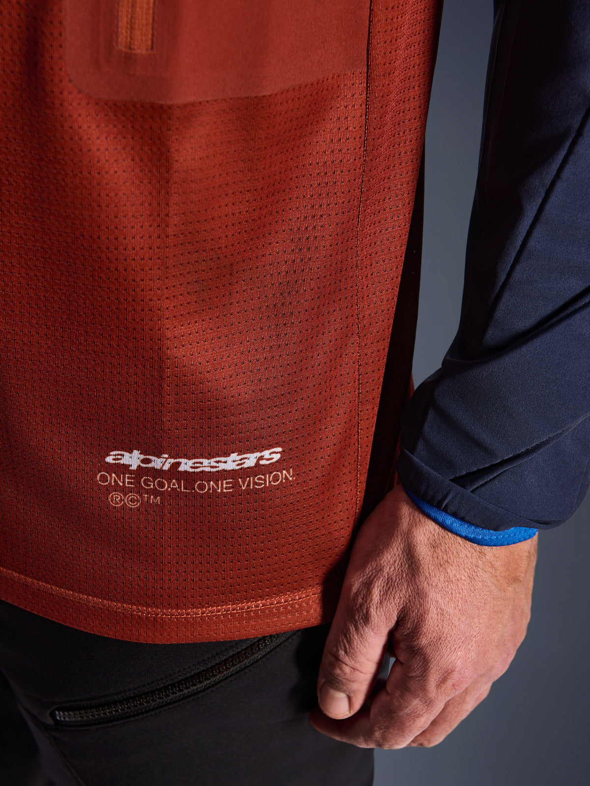 Alpinestars Maxdura Dual Jersey, maillot de moto aventure, brun rouille et bleu Mood Indigo, vue rapprochée montrant le tissu en maille du torse et la manche tissée bleu marine, logo Alpinestars blanc « One Goal One Vision » sur l'ourlet inférieur.