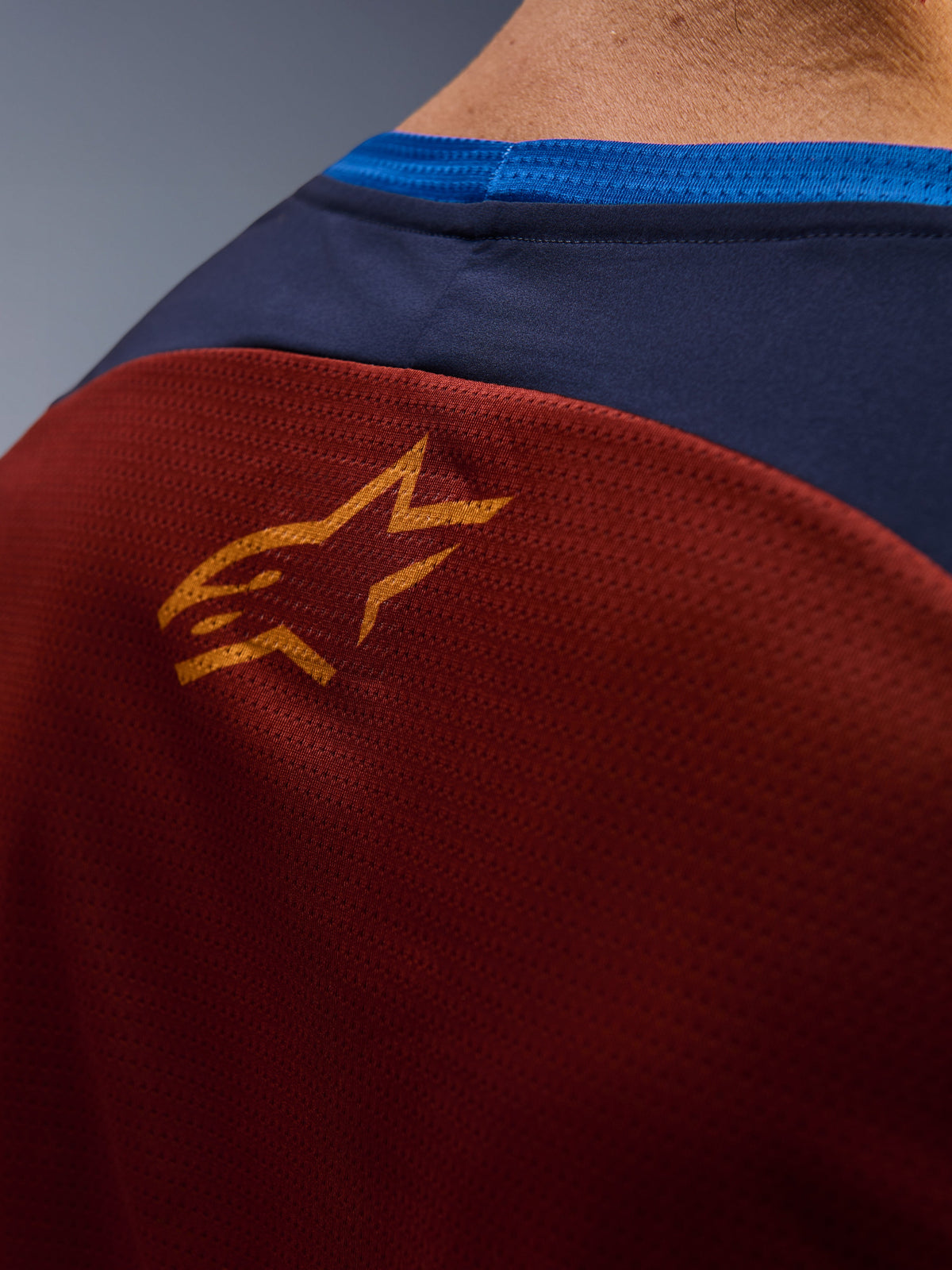 Gros plan du maillot Alpinestars Maxdura Dual, tissu en maille marron rouille au dos, petit logo étoile orange, empiècement supérieur bleu foncé et bordure du col bleu vif, vue détaillée d'un maillot de moto d'aventure