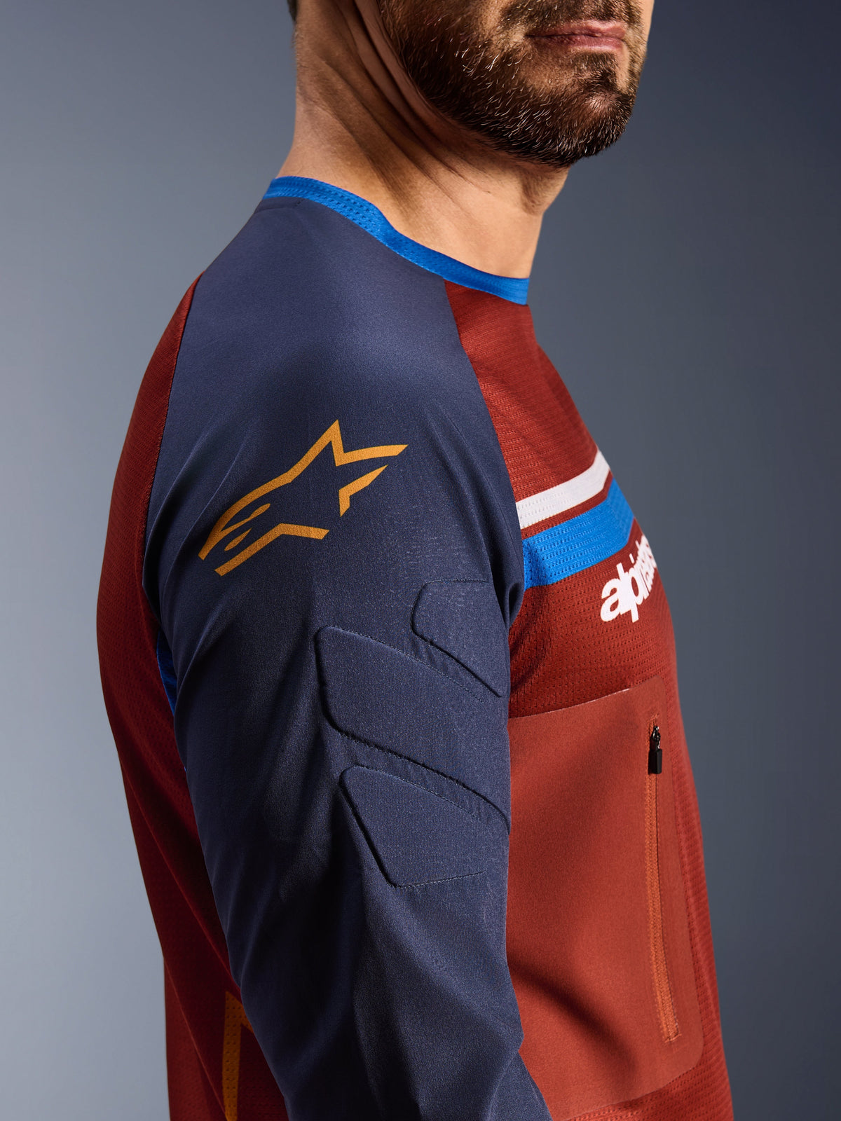 Maillot Alpinestars Maxdura Dual, Brun Rouille et Indigo, vue de profil montrant les manches rembourrées avec le logo étoile jaune, construction du torse en maille avec poche poitrine zippée, vêtement de moto technique pour la conduite aventure