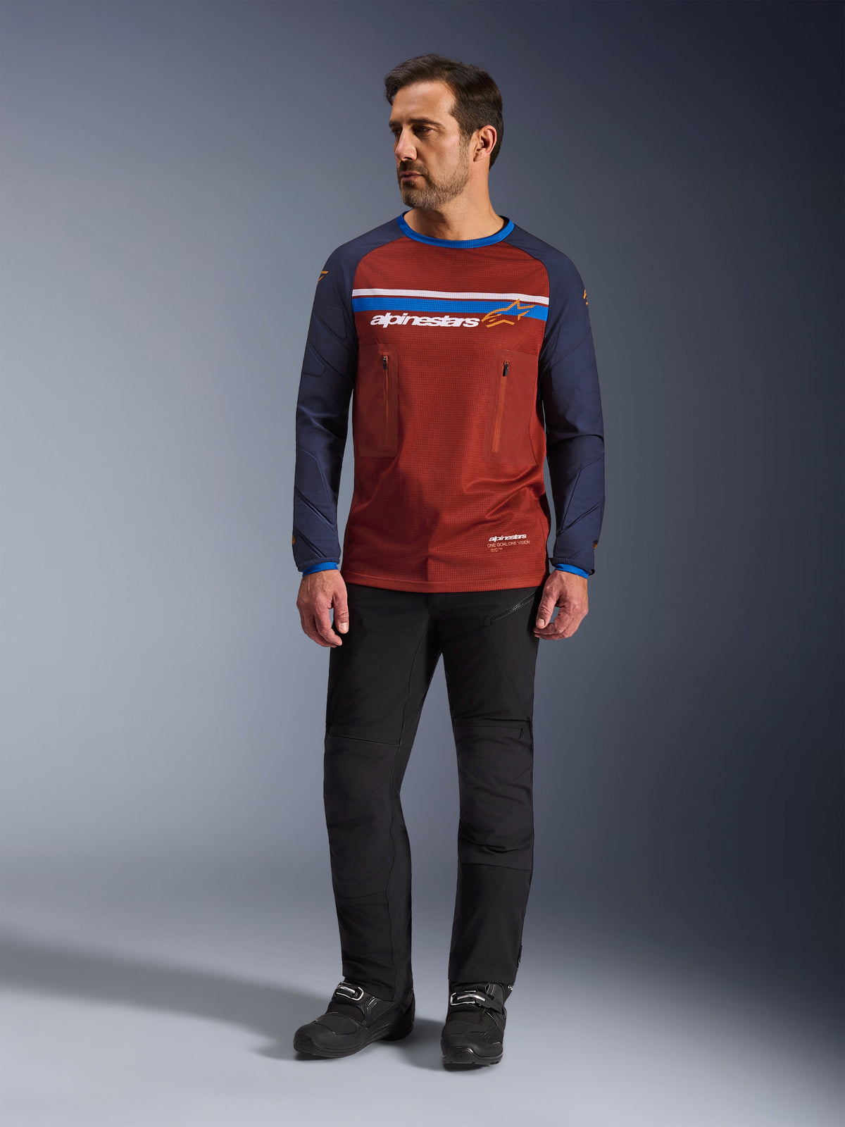 Maillot Alpinestars Maxdura Dual, brun rouille et bleu Mood Indigo, maillot de moto d'aventure avec torse en maille et manches bleu marine, porté par un mannequin avec un pantalon de moto noir et des bottes noires, vêtement technique dual-sport pour la conduite tout-terrain
