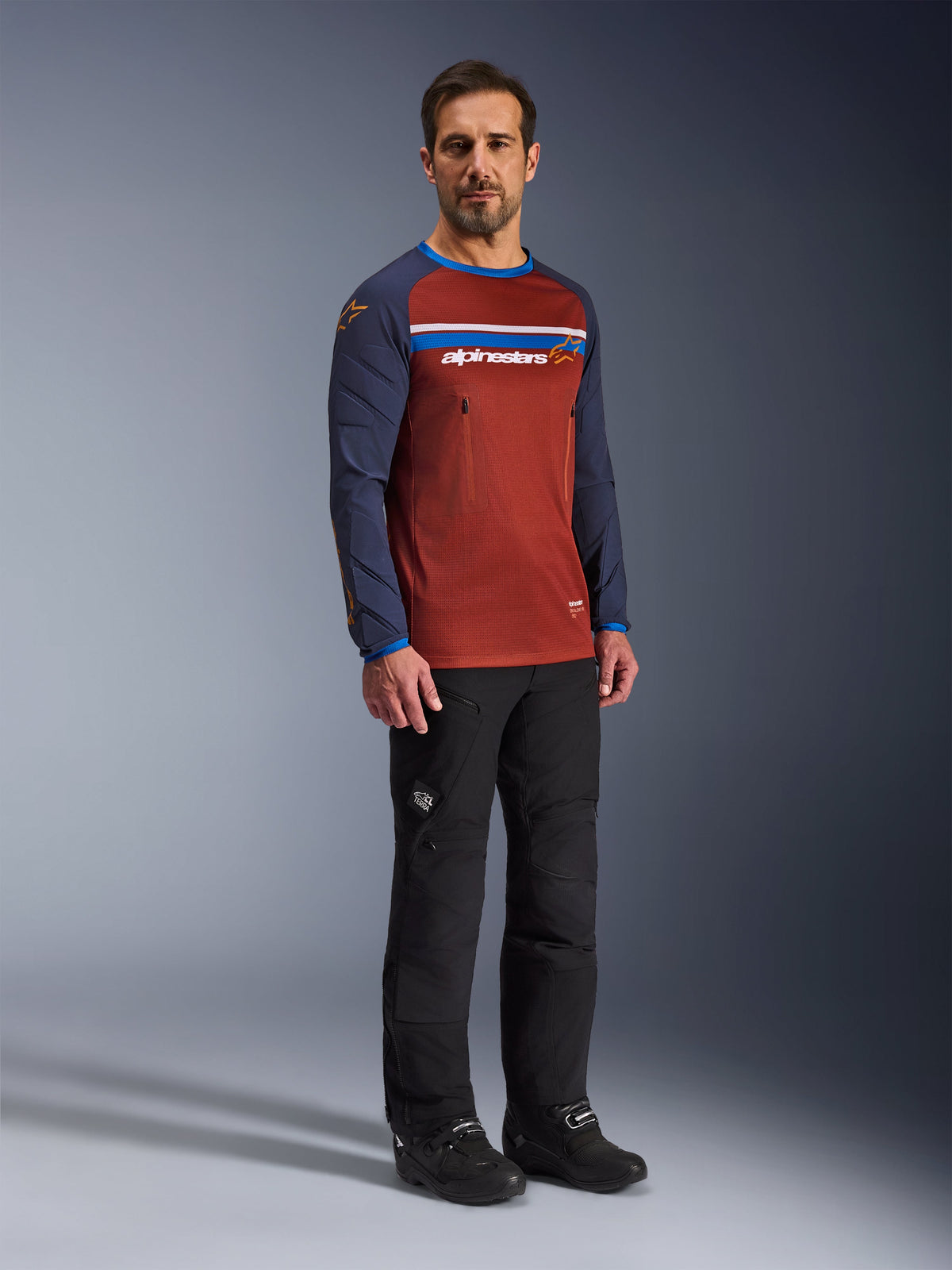 Homme portant le maillot Alpinestars Maxdura Dual en Rust Brown et Mood Indigo, avec torse en maille et manches rembourrées, associé à un pantalon de moto et des bottes noirs, équipement de moto adventure