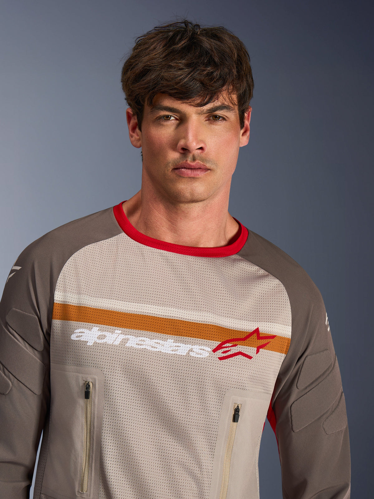 Mannelijk model draagt de Alpinestars Maxdura Dual Jersey in Aluminum Walnut, adventure motorjersey met een torso van volledig mesh, slijtvaste mouwen, rode kraag en borstzakken met rits, voorzien van oranje en witte borststrepen met Alpinestars-logo.