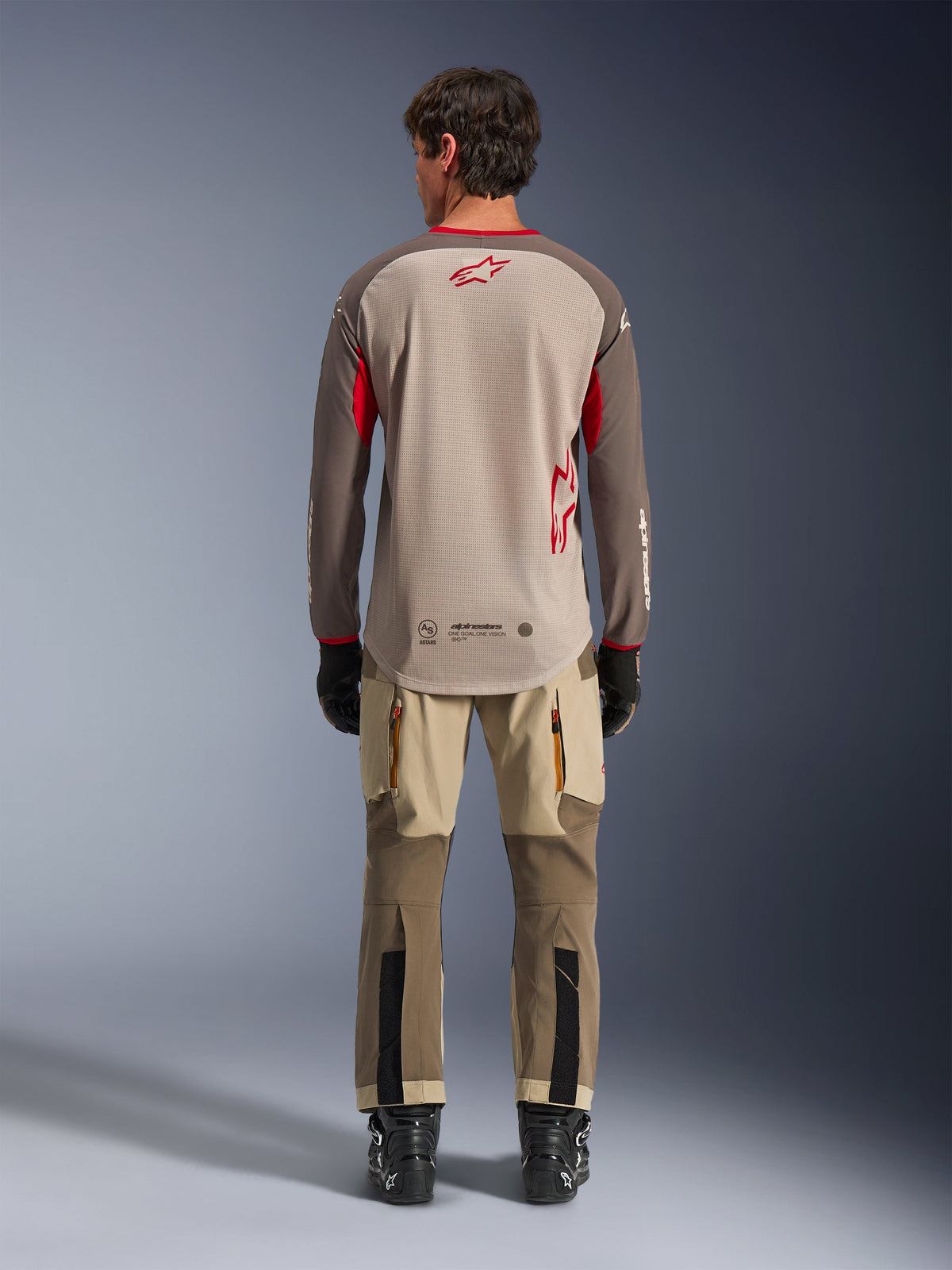 Alpinestars Maxdura Dual Jersey in Aluminum Walnut, achteraanzicht, beige mesh torso met bruine mouwen en rode logo's, verlaagde achterzoom, gecombineerd met adventure motorbroek en zwarte laarzen.