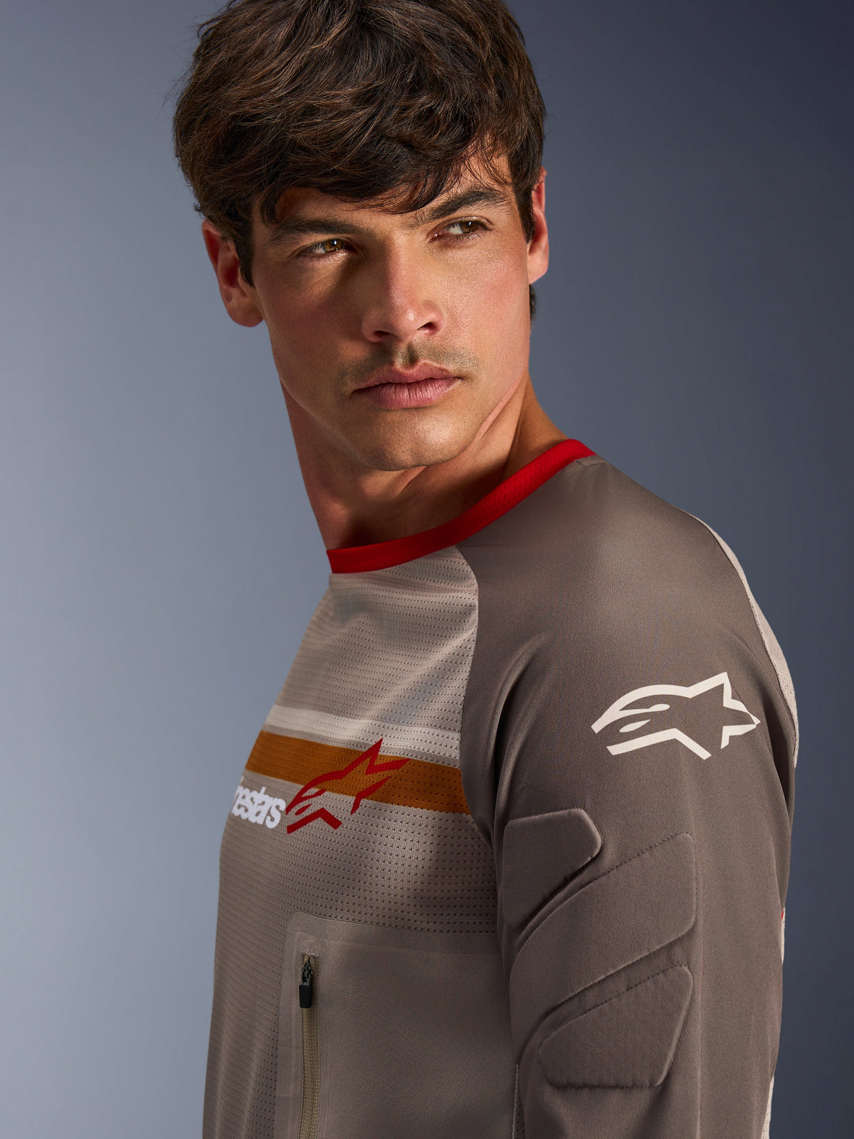 Mannelijk model met Alpinestars Maxdura Dual Jersey in Aluminum Walnut, met een geventileerde mesh torso, rode kraag en oranje borststreep. Close-up weergave van slijtvaste mouw met geïntegreerde padding en een borstzak met ritssluiting voor adventure rijden.