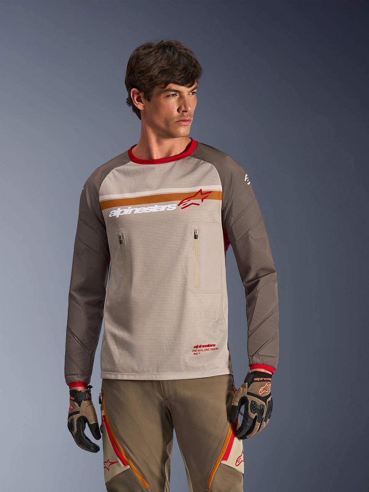 Alpinestars Maxdura Dual Jersey in Aluminum Walnut, adventure motorjersey met een ademende mesh torso, walnootbruine slijtvaste mouwen en borstzakken met ritssluiting, gedragen door een model met bijpassende off-road uitrusting