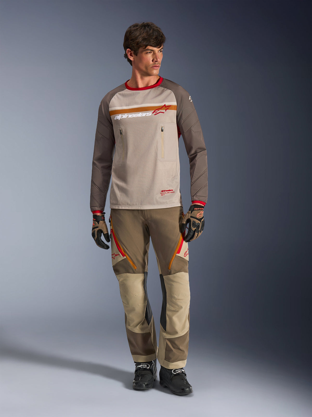 Alpinestars Maxdura Dual Jersey in Aluminum Walnut, adventure motorjersey met zandkleurige mesh torso en bruine mouwen, rode kraag en horizontale borststreep met Alpinestars-logo, gedragen met bijpassende adventure broek en handschoenen.