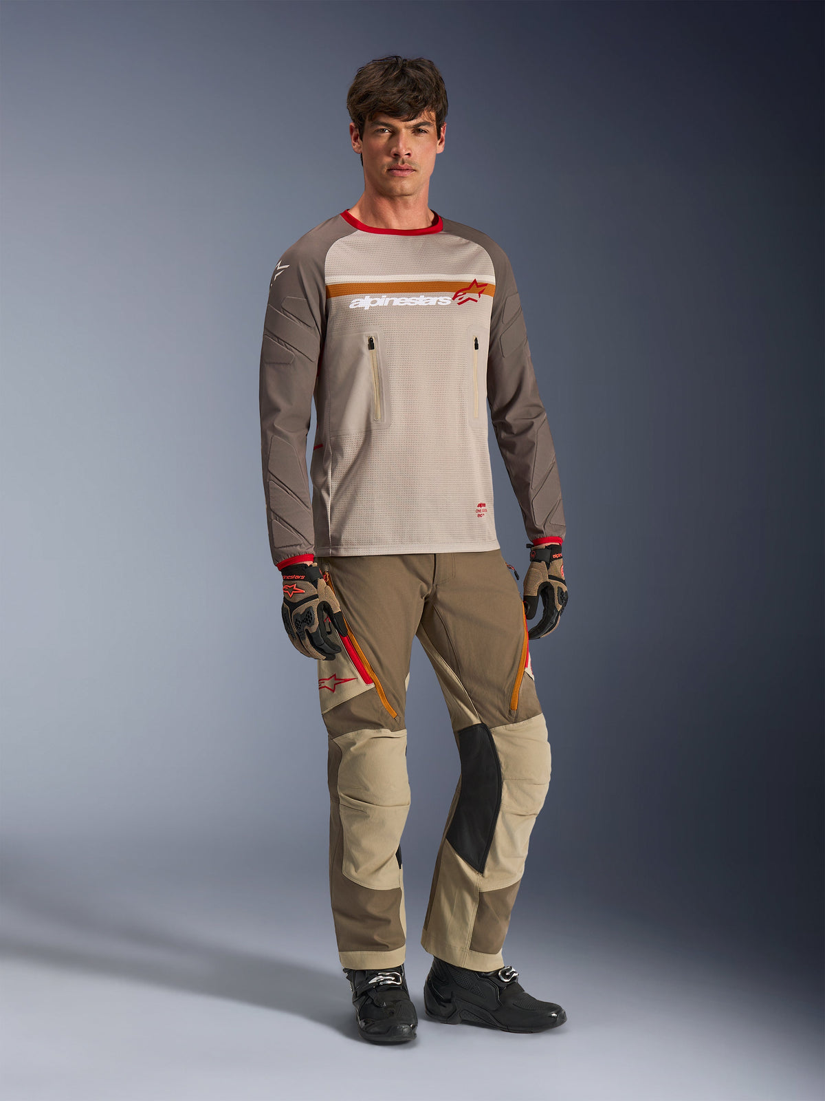 Model draagt de Alpinestars Maxdura Dual Jersey in Aluminum Walnut, voorzien van een grijze mesh torso, bruine mouwen met rode boorden en borstzakken met ritssluiting, gecombineerd met bijpassende off-road uitrusting voor adventure-rijden.
